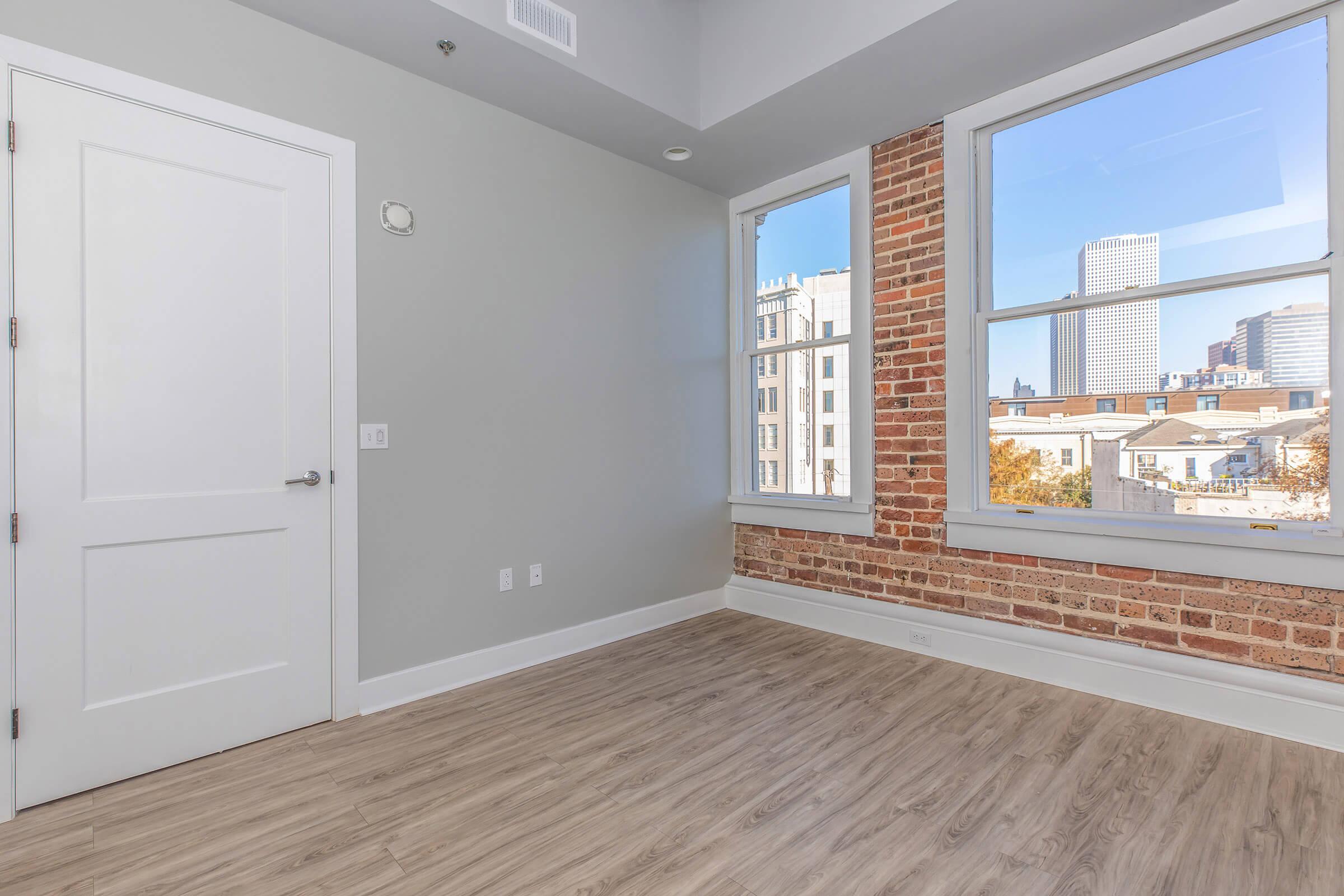 Bright, modern room featuring light gray walls, large windows with a view of a city skyline, and exposed brick accents. The floor is a light wood finish, and the space includes a white door and minimalist design details for an inviting atmosphere.