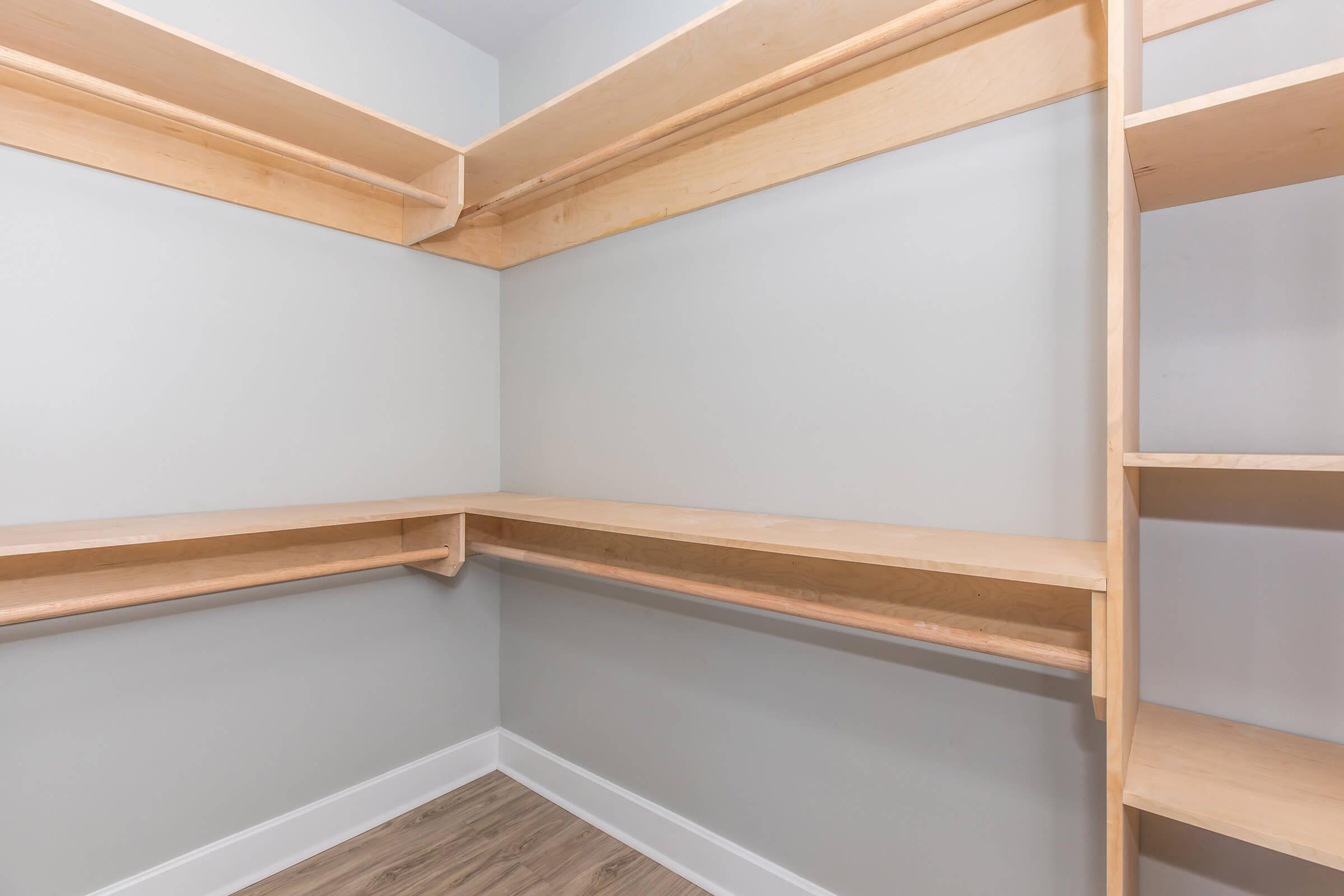 A minimalist walk-in closet with light-colored wooden shelving and a neutral-colored wall. Two long shelves run horizontally along the upper wall, with a lower shelf corner table positioned against one wall. The flooring is a light wood finish, providing a spacious and organized appearance.