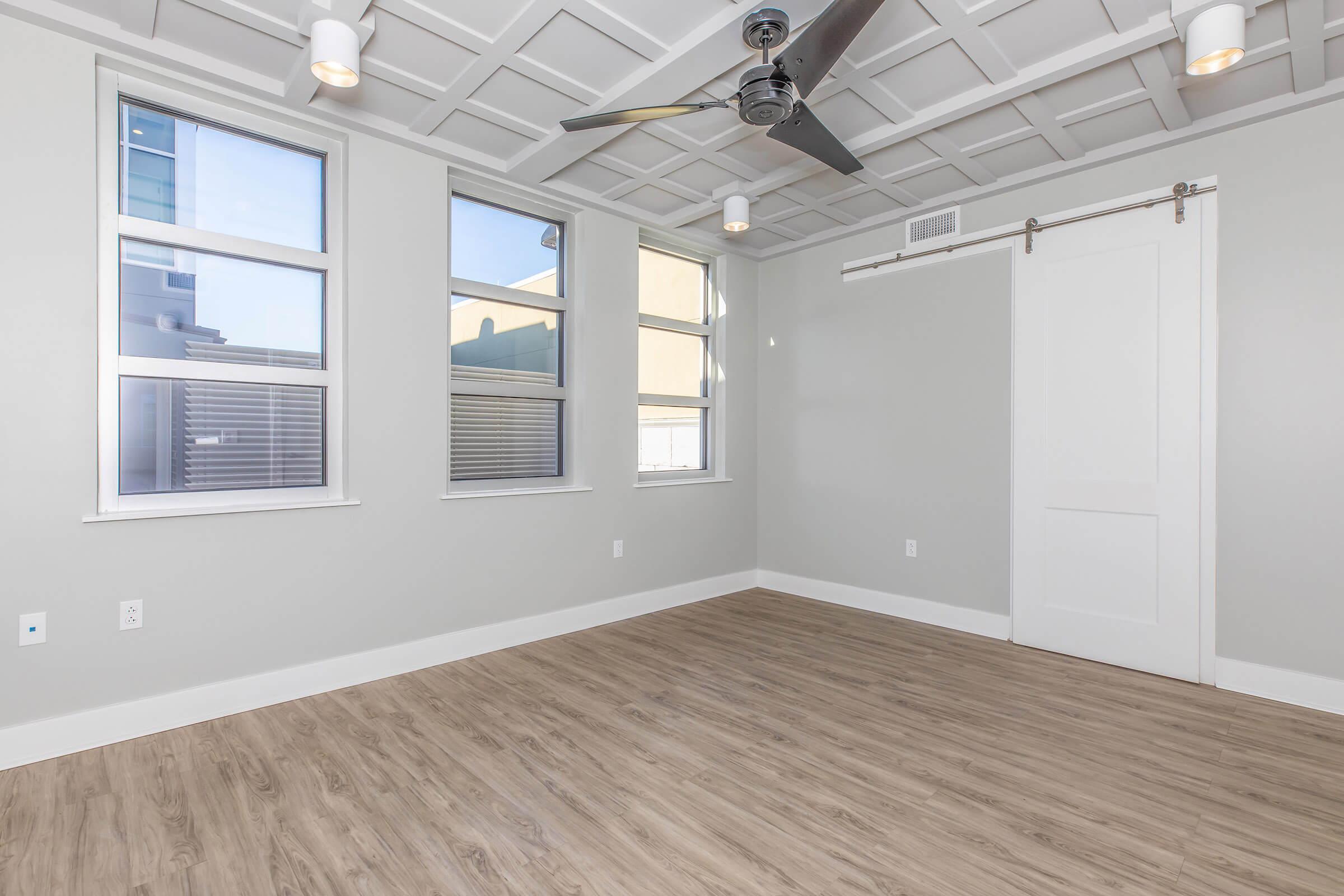 A well-lit empty room featuring light gray walls, large windows, and wooden flooring. The ceiling has a modern design with a ceiling fan in the center. One wall has a sliding door, and the space is structured to allow for natural light and a minimalist aesthetic. No furniture is present.