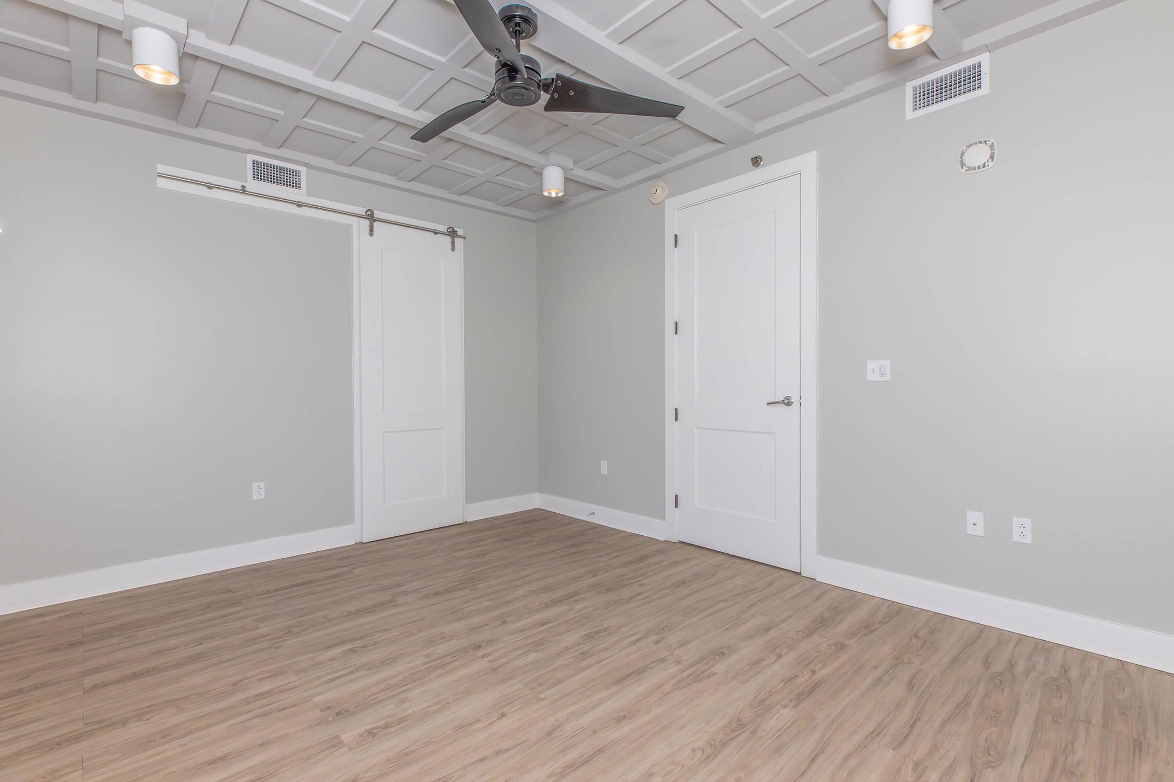 A spacious, empty room with light gray walls and wooden flooring. The room features a ceiling fan, recessed lighting, and a sliding barn door. A standard white door is visible on one wall, while the opposite wall has a small vent. The overall design is modern and minimalist.