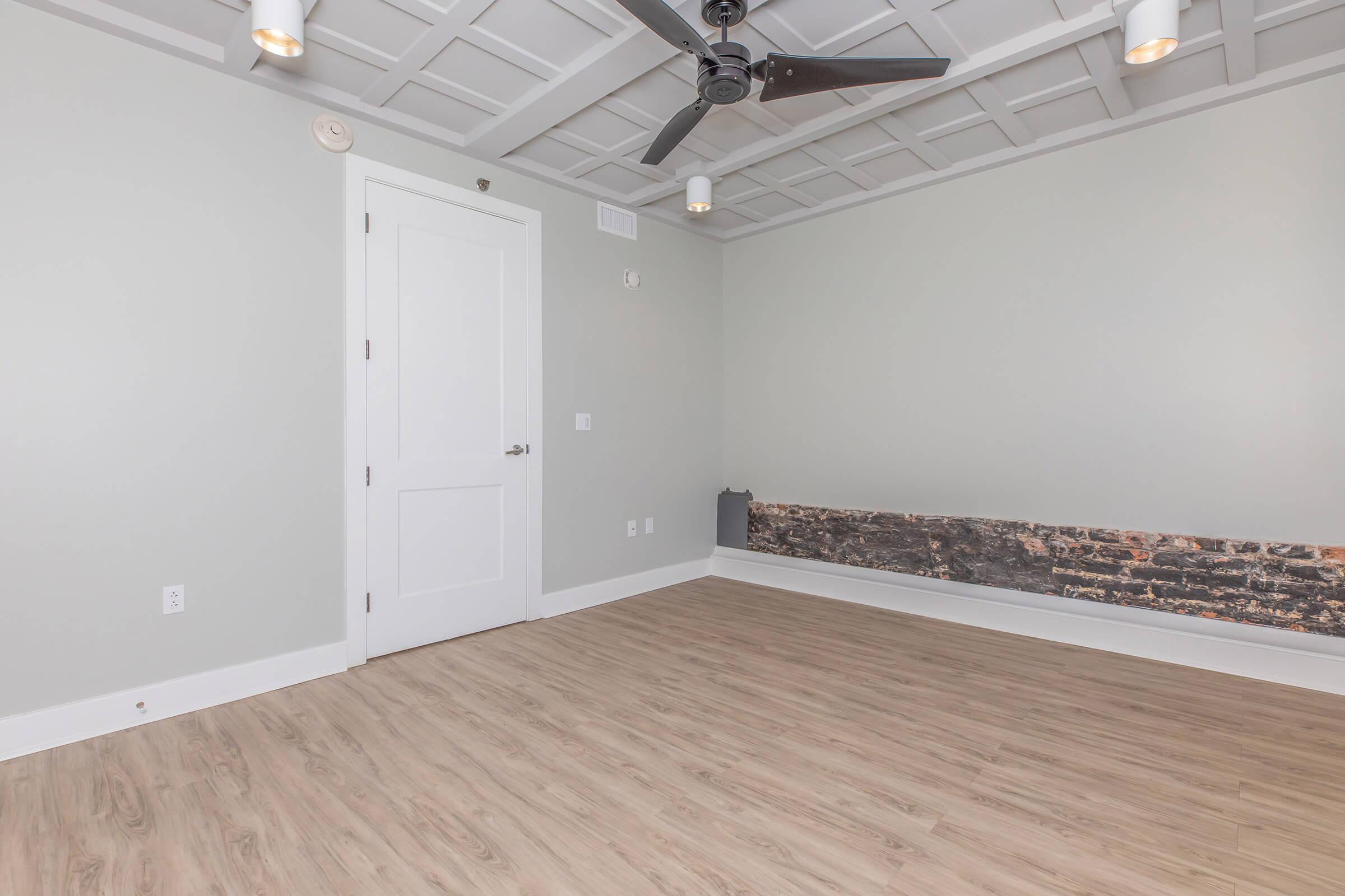 A spacious, modern room with light gray walls and a ceiling featuring decorative panels. The floor is made of light wood, and the room includes a ceiling fan. There's a white door and a section of exposed brick along the lower wall, adding character to the otherwise minimalistic design.