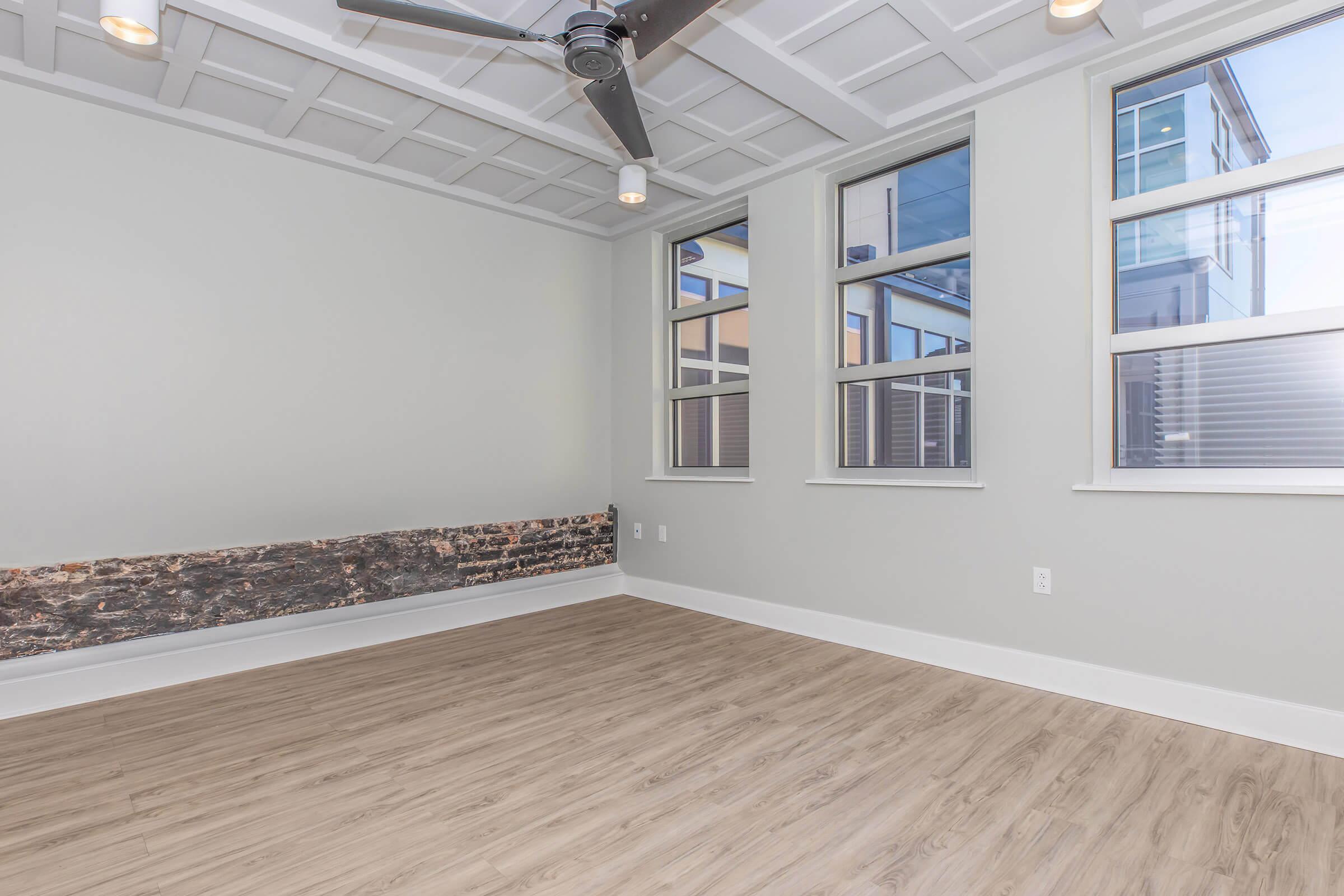 A bright, empty room featuring light-colored walls, wood-like flooring, and a modern ceiling fan. Large windows allow natural light to enter, while a textured stone wall adds visual interest along one side. The space has a minimalist design and is ready for decorating.