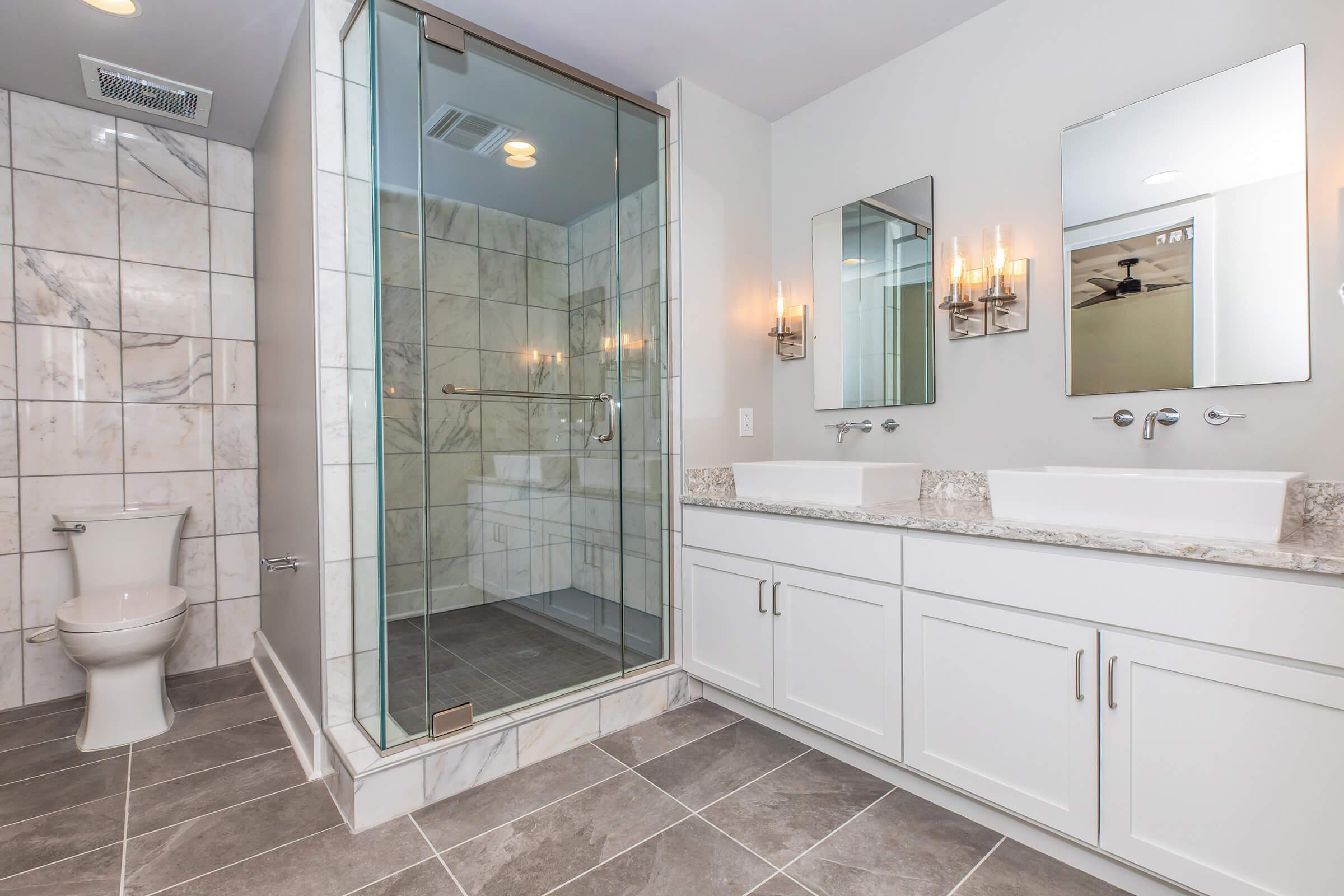 Modern bathroom featuring a glass-enclosed shower with marble tiles, a double vanity with marble countertops and two sinks, wall-mounted mirrors, stylish sconces, and a toilet. The floor is tiled with large gray tiles, and the overall decor is sleek and contemporary.