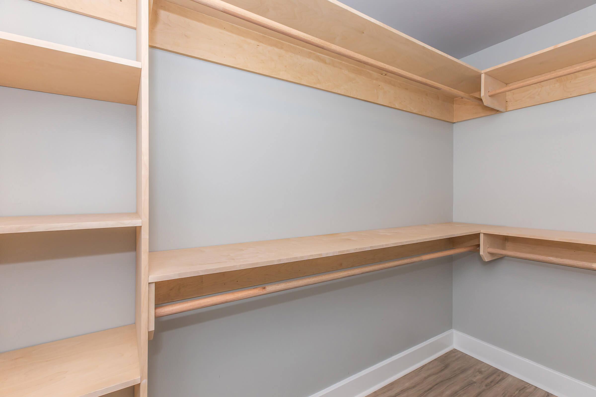 Empty closet with wooden shelves and a hanging rod, featuring light-colored wood against a pale gray wall and a light-colored floor. The design is minimalistic and modern, providing ample space for storage.