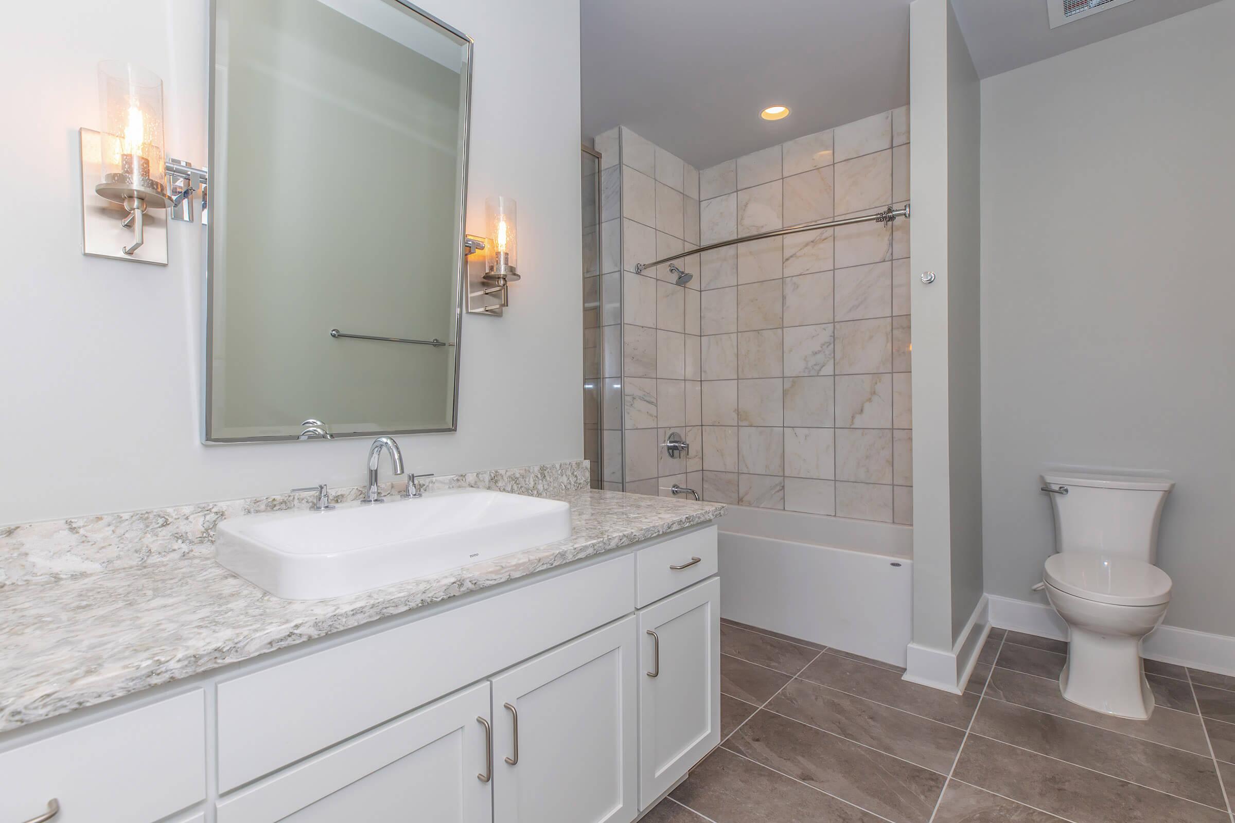 A modern, well-lit bathroom featuring a white vanity with a rectangular sink, a large mirror, and two wall sconces. The tiled wall and shower area have a glass door, while a white bathtub is adjacent. The flooring is a neutral tile, and a white toilet is placed in the corner.