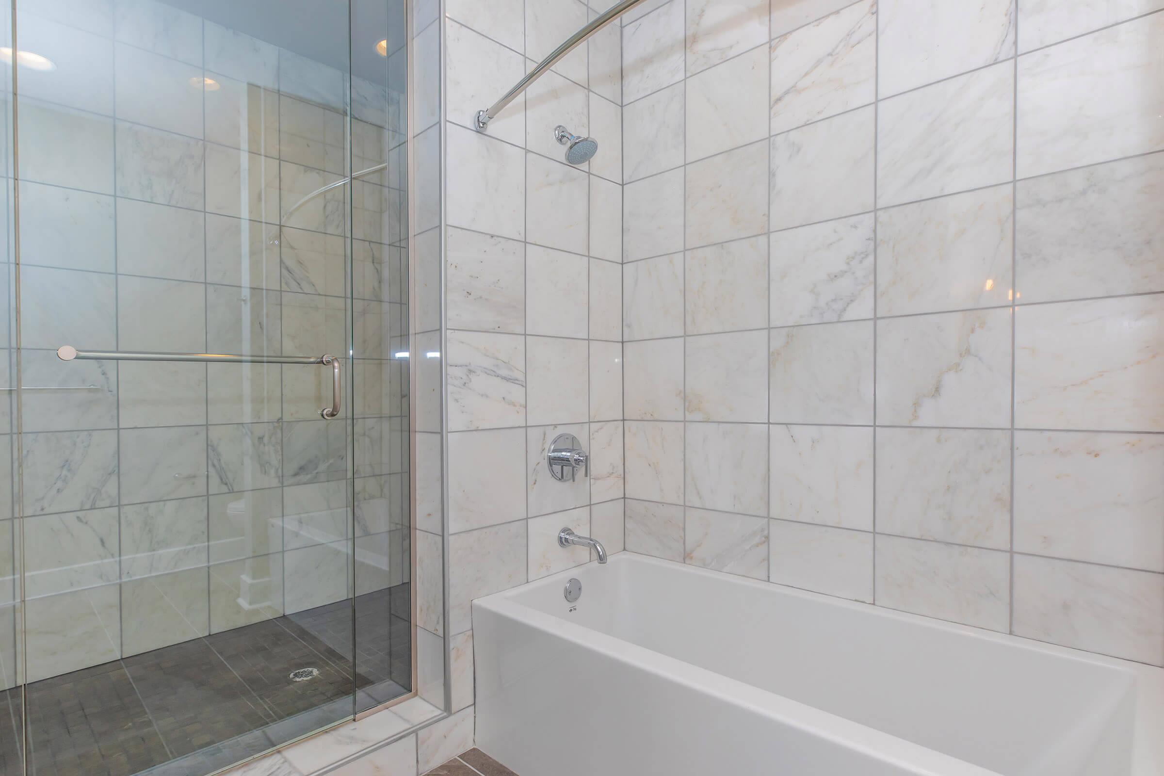 A modern bathroom featuring a glass shower enclosure with tiled walls and a bathtub. The walls are covered in light-colored marble tiles, and the floor has dark wood-style tiles. The space is well-lit with natural light, creating a clean and spacious appearance.