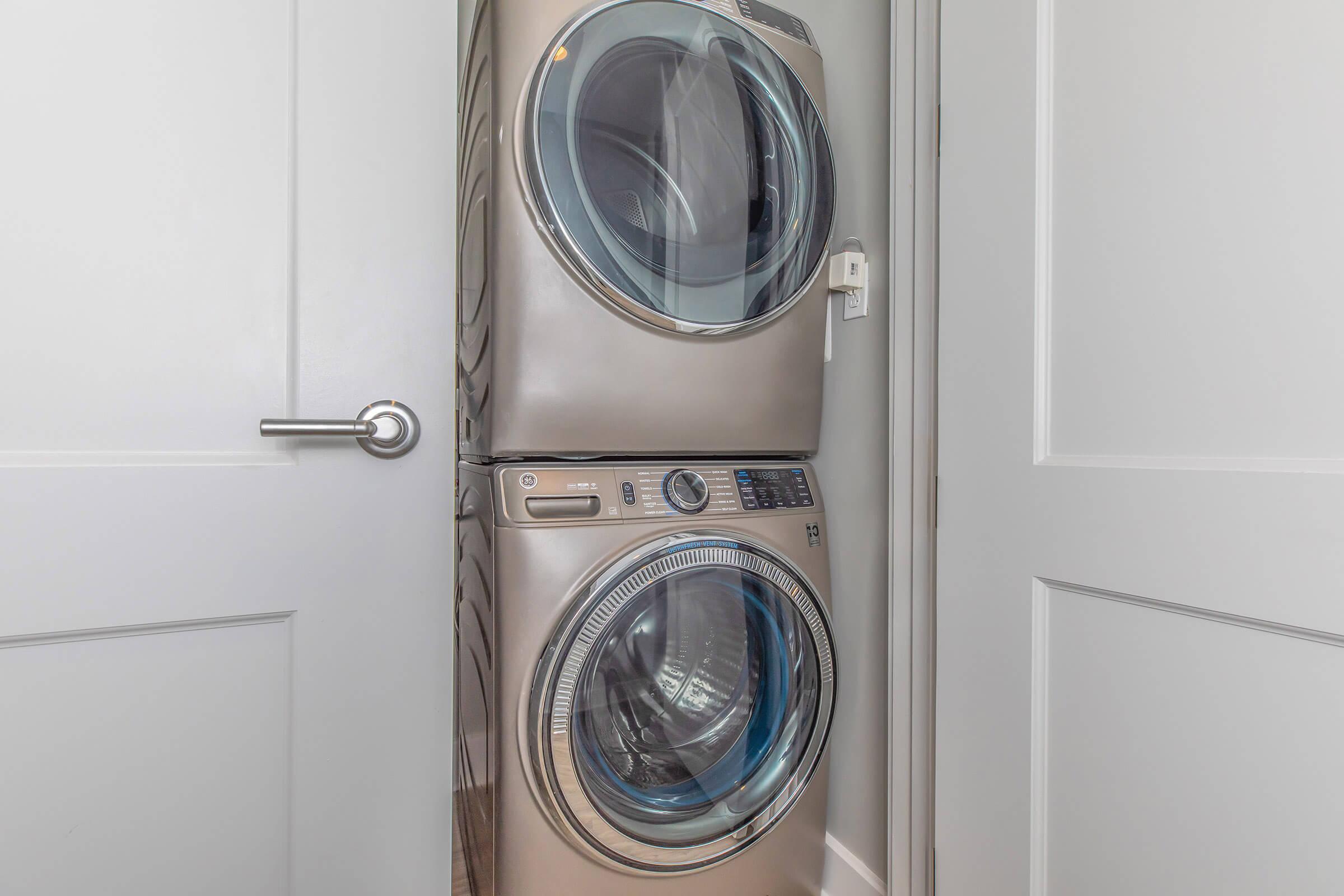 A stackable washer and dryer in a small laundry space with a light gray wall and a white door partially opened. The appliances have a modern design with shiny metallic finishes and clear circular doors.