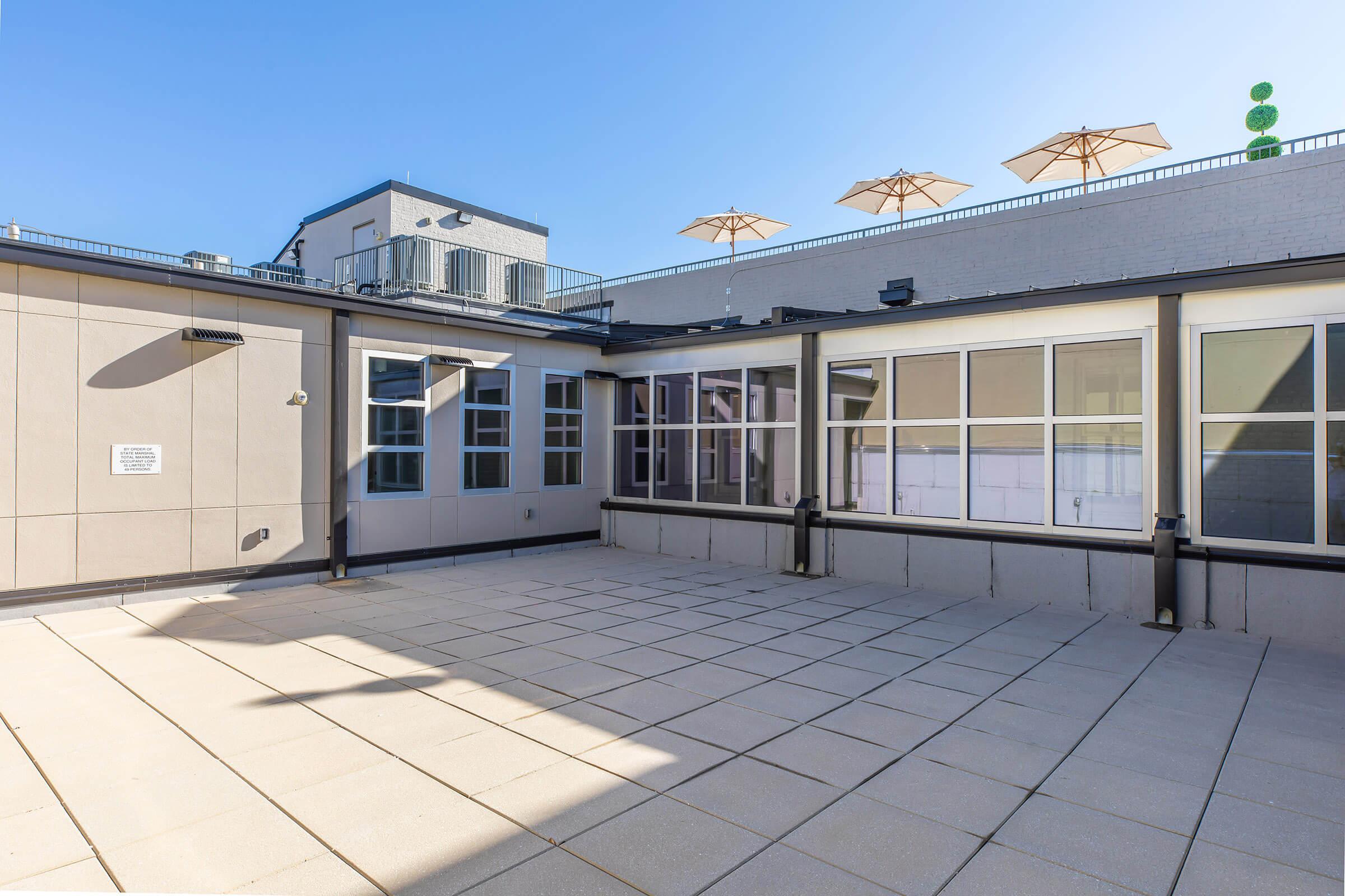 A large open rooftop space with a flat, tiled floor, featuring several light-colored umbrellas set up for shade. The sky is clear and blue, and there are large windows on one side of the rooftop. The area appears well-lit and inviting, suitable for gatherings or relaxation.