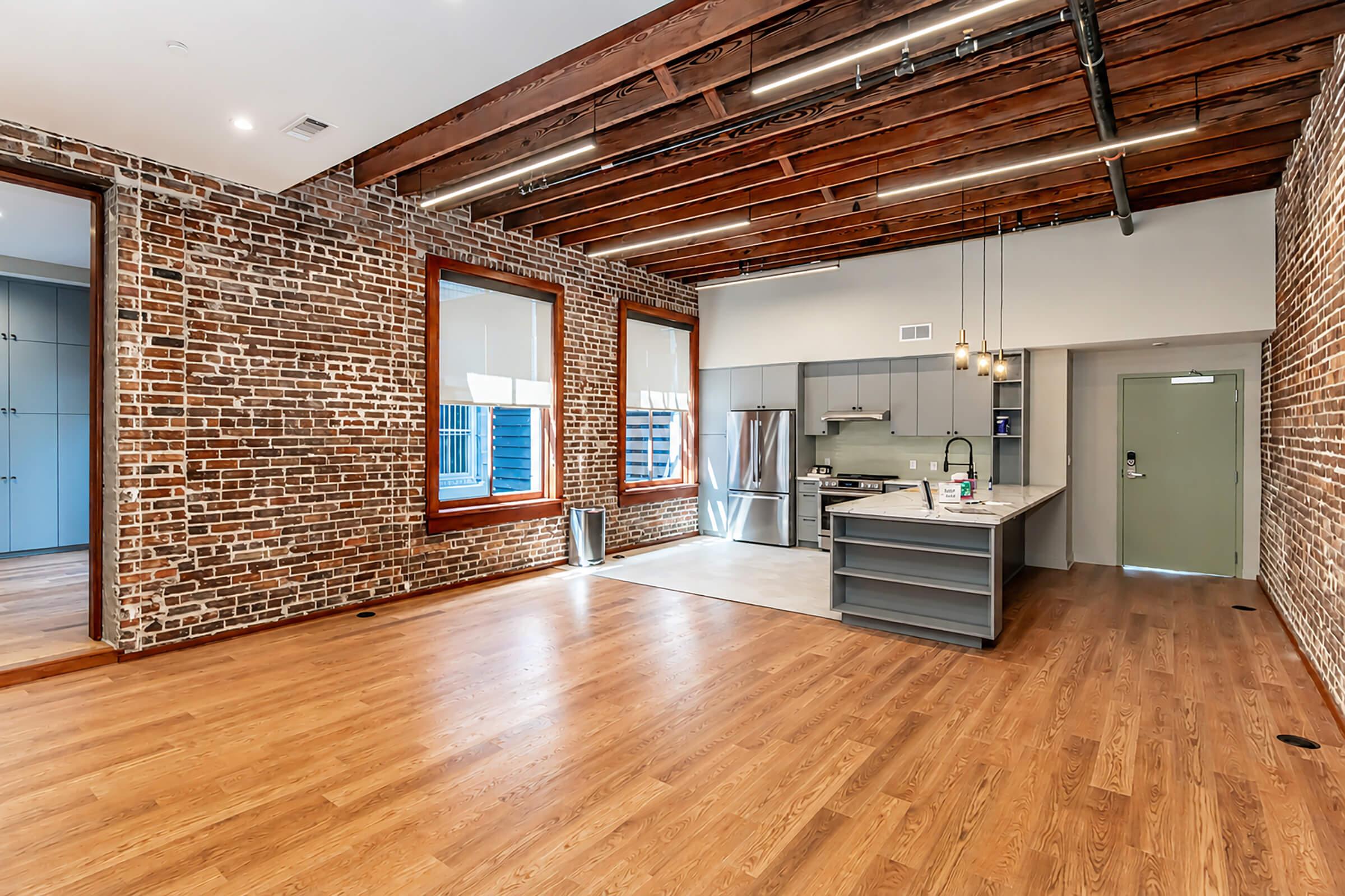 Open-concept living space with exposed brick walls, hardwood floors, and large windows allowing natural light. Features a modern kitchen with stainless steel appliances and a sleek island. Contemporary design elements are visible, along with a door leading to a hallway.