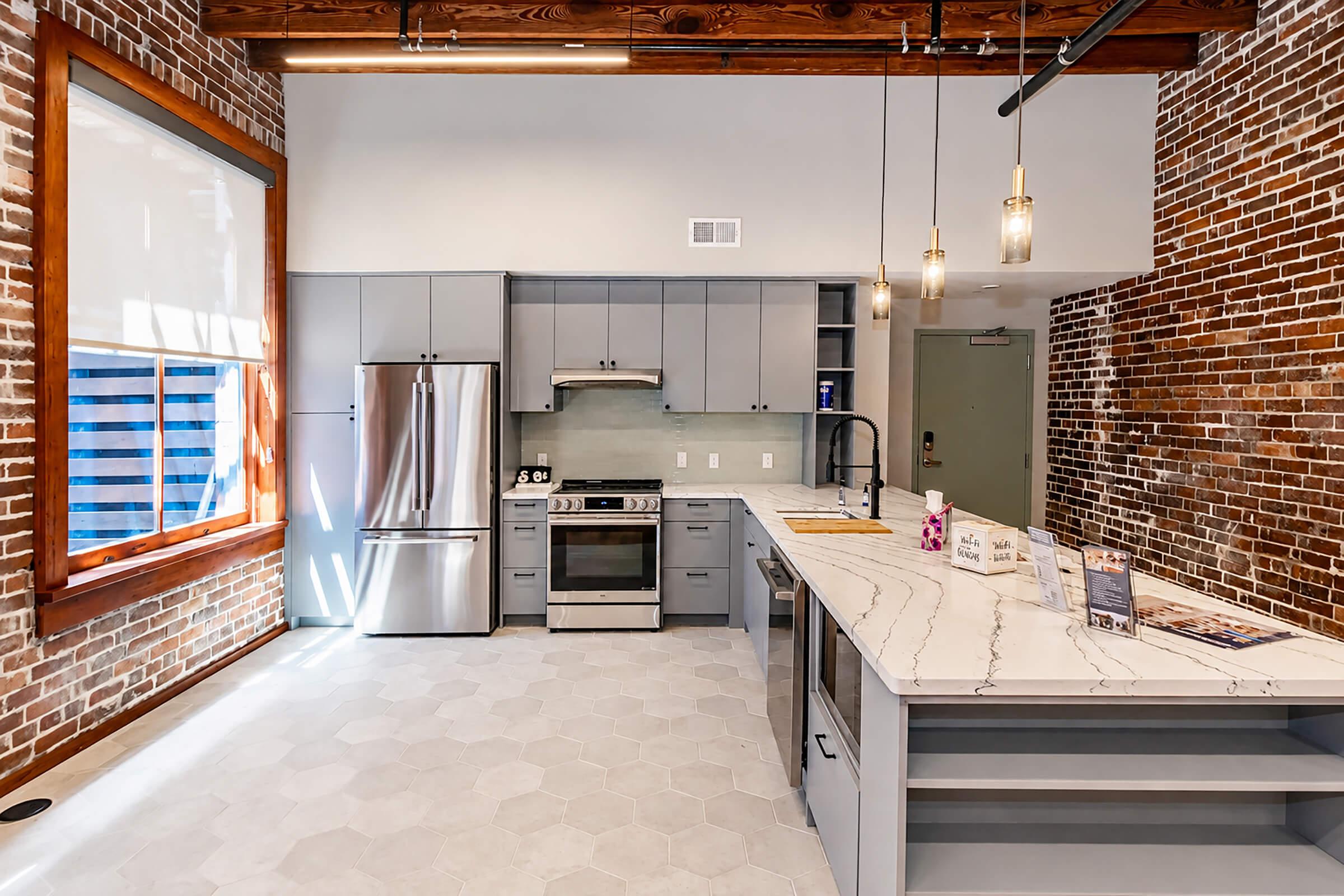 Modern kitchen featuring gray cabinets, stainless steel appliances, and a marble countertop. Exposed brick walls add an industrial touch. The space has large windows with natural light, hexagonal tile flooring, and stylish pendant lighting above the island.