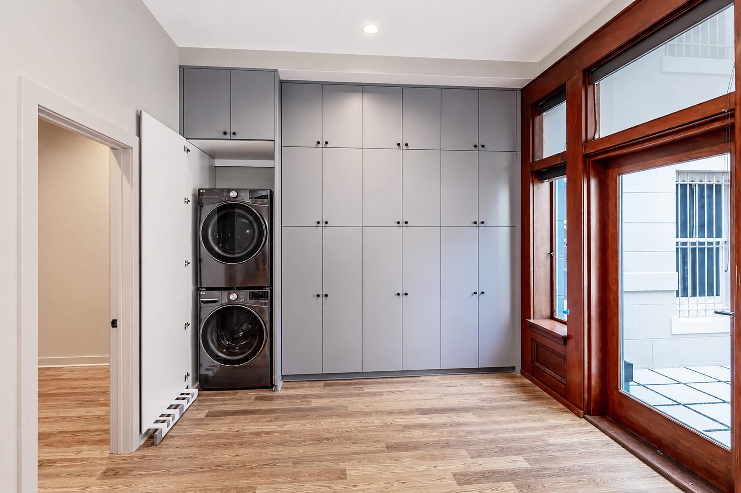 A modern laundry room featuring stacked washing and drying machines against a wall with gray cabinetry. The space has hardwood flooring and large windows with wooden frames, allowing natural light to fill the room. The design is clean and minimalistic.
