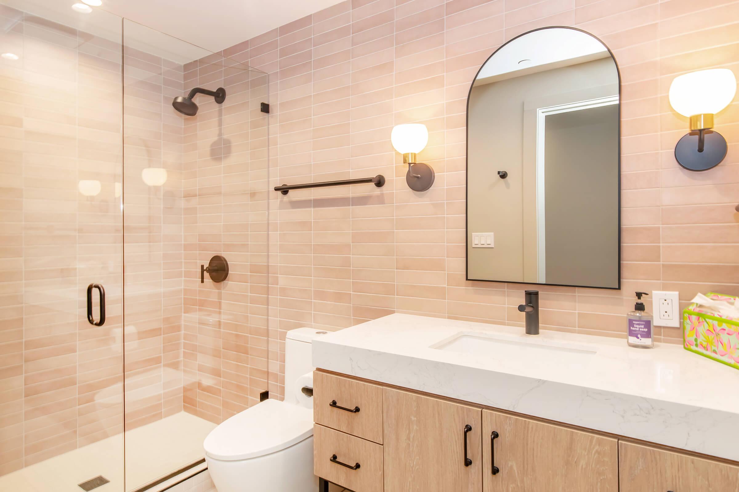 A modern bathroom featuring a glass shower enclosure, a rectangular mirror with an arch top, a wall-mounted light fixture, and a light wood vanity with a white marble countertop. The walls are adorned with soft pink tiles, and a toilet is visible beside the vanity.