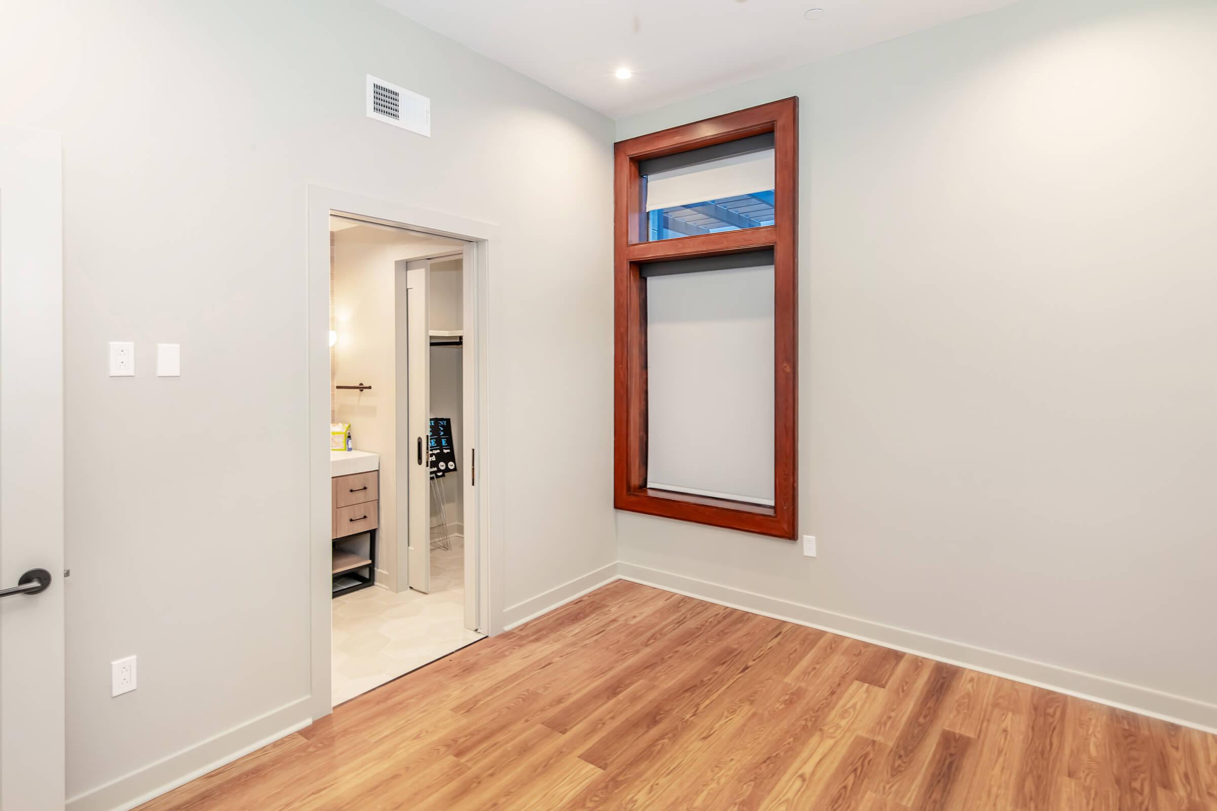 A modern, minimalistic room with light gray walls and hardwood flooring. On the left, a doorway leads to a bathroom, partially visible. The right side features a large window with dark wooden trim, allowing natural light to enter the space. The overall aesthetic is clean and contemporary.