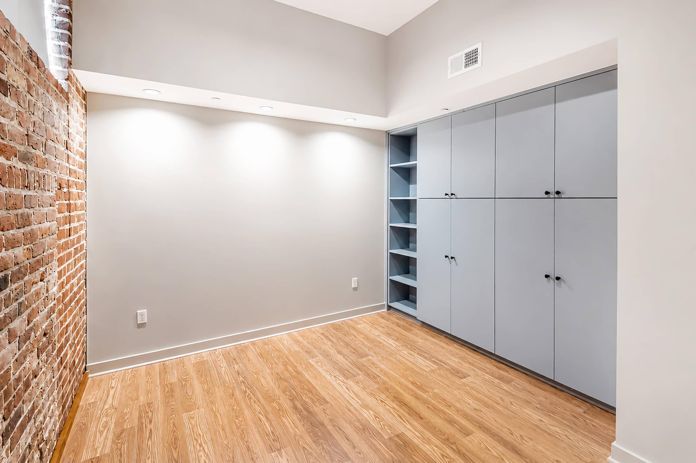 A modern, minimalist room featuring a combination of exposed brick and gray walls. There are built-in cabinets with a smooth finish on one side, and the floor is made of polished wood. Recessed lighting illuminates the space, creating a bright atmosphere. The overall design is sleek and contemporary.