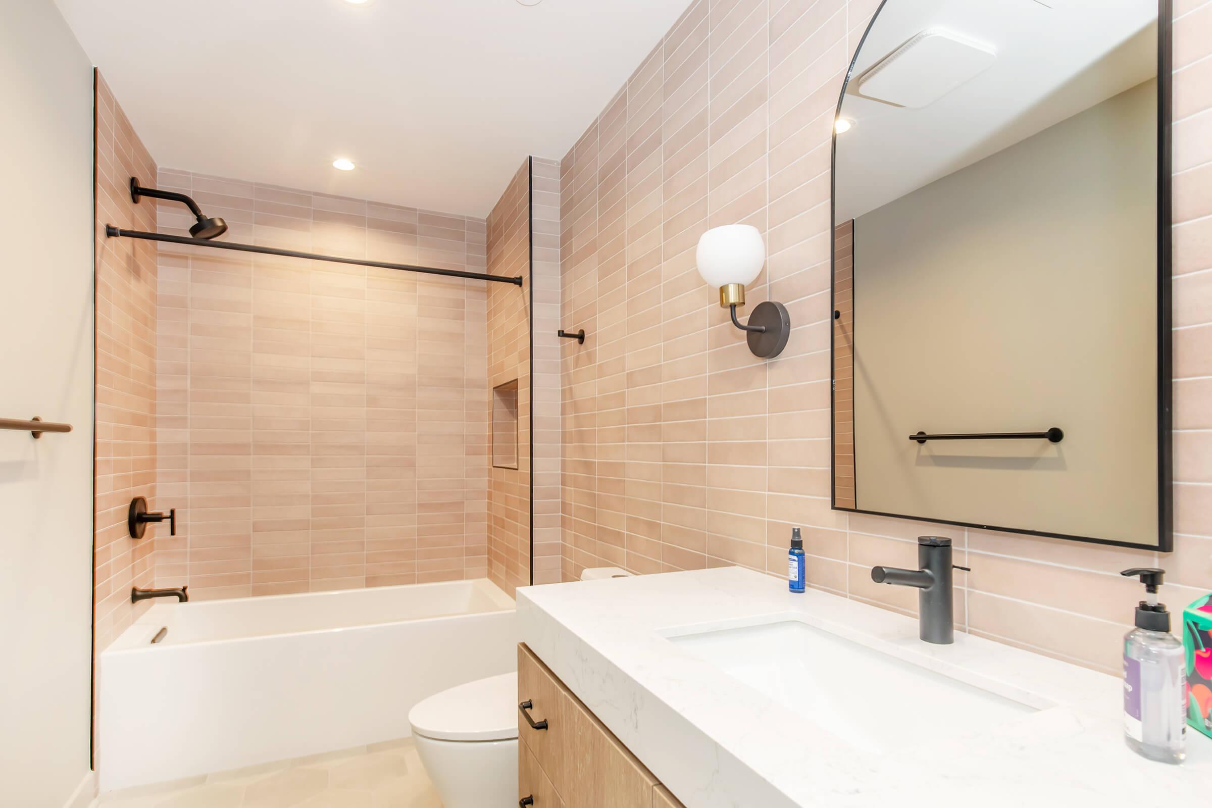 A modern bathroom featuring a light-colored tile design. It includes a bathtub with a shower, a sleek sink with a marble countertop, a large mirror, and wall-mounted fixtures. The space is well-lit, highlighting the neutral tones and contemporary style.