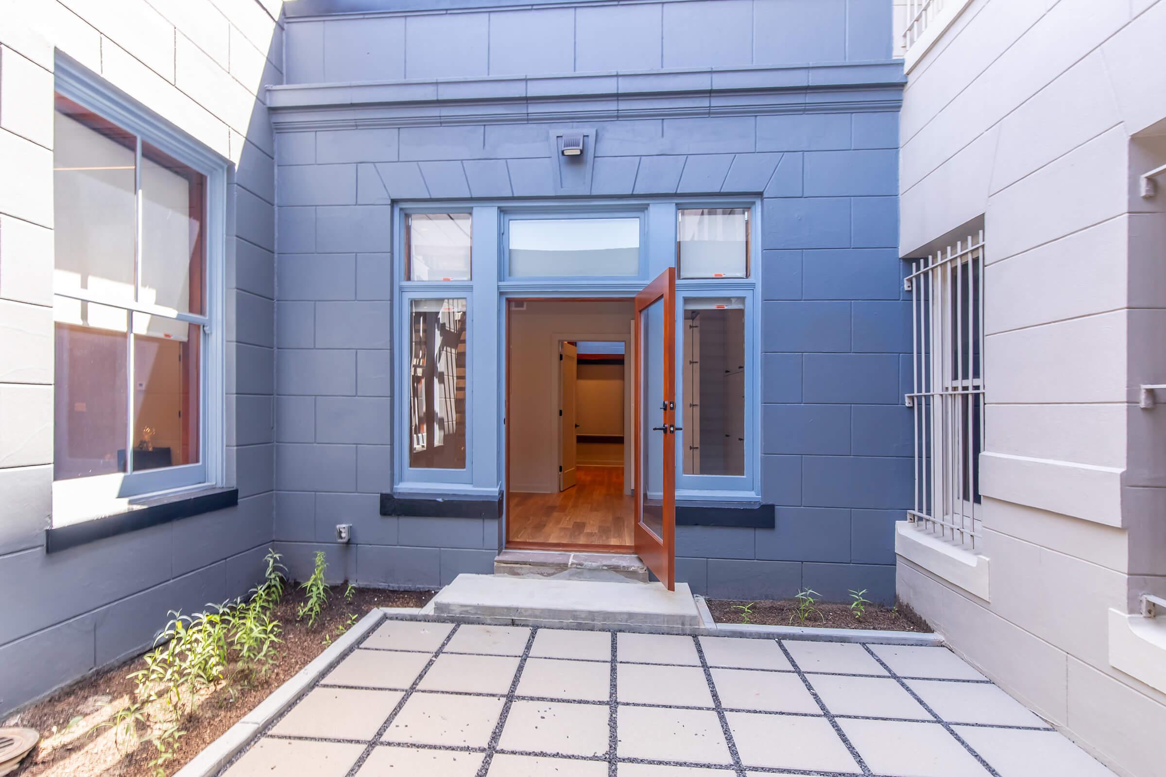 A small courtyard with a paved pathway leading to an open door. The entrance features a painted blue wall and large windows. Inside, a glimpse of wooden flooring is visible, suggesting a spacious interior. Surrounding the pathway are small plants and well-maintained stone edges.