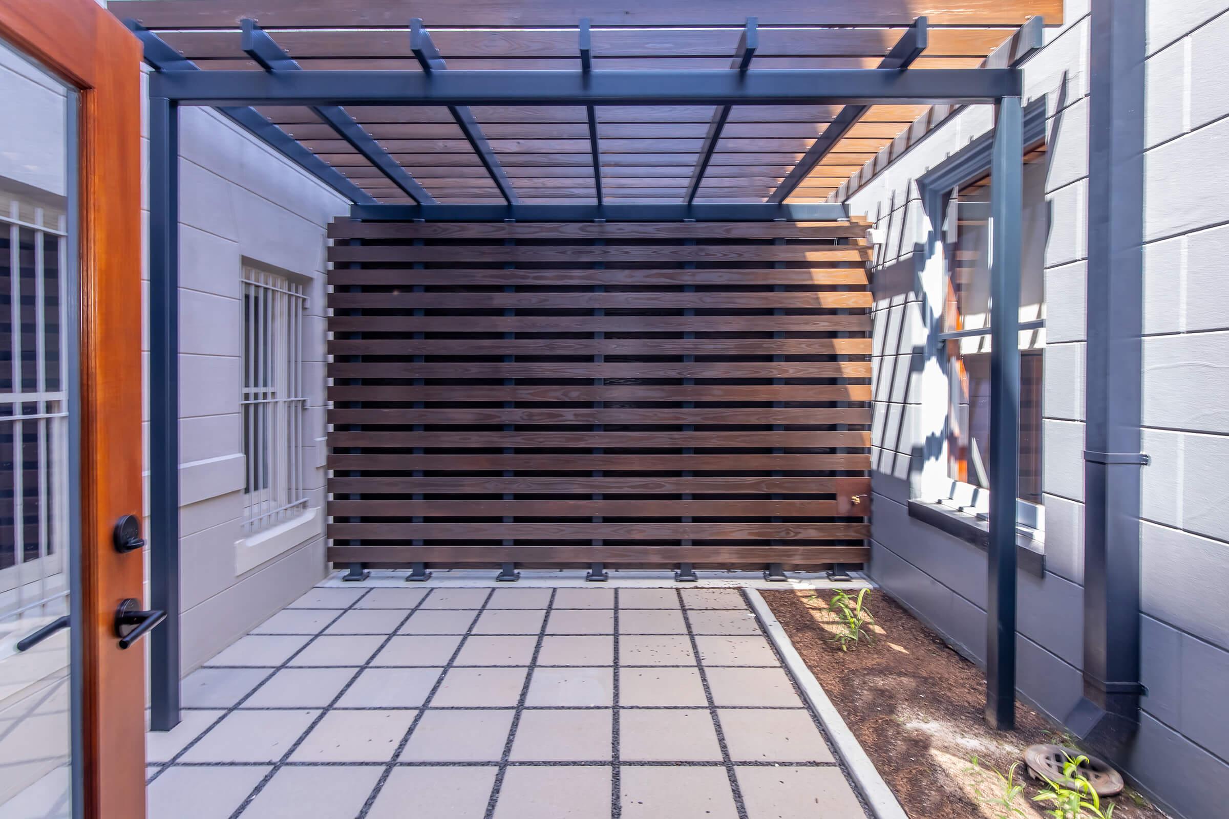A modern outdoor space featuring a wooden pergola, a slatted wooden wall, and a tiled floor. The area is well-lit, showcasing neat landscaping along the edges, creating a serene atmosphere suitable for relaxation or gatherings.