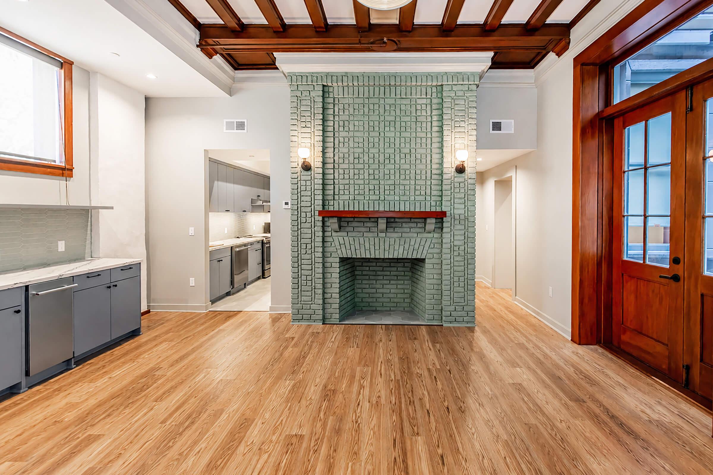 A spacious room featuring wooden beams on the ceiling, light-colored walls, and a distinctive green tiled fireplace. The flooring is light wood, and there are large windows allowing natural light. The kitchen area is partially visible with gray cabinets and modern appliances.