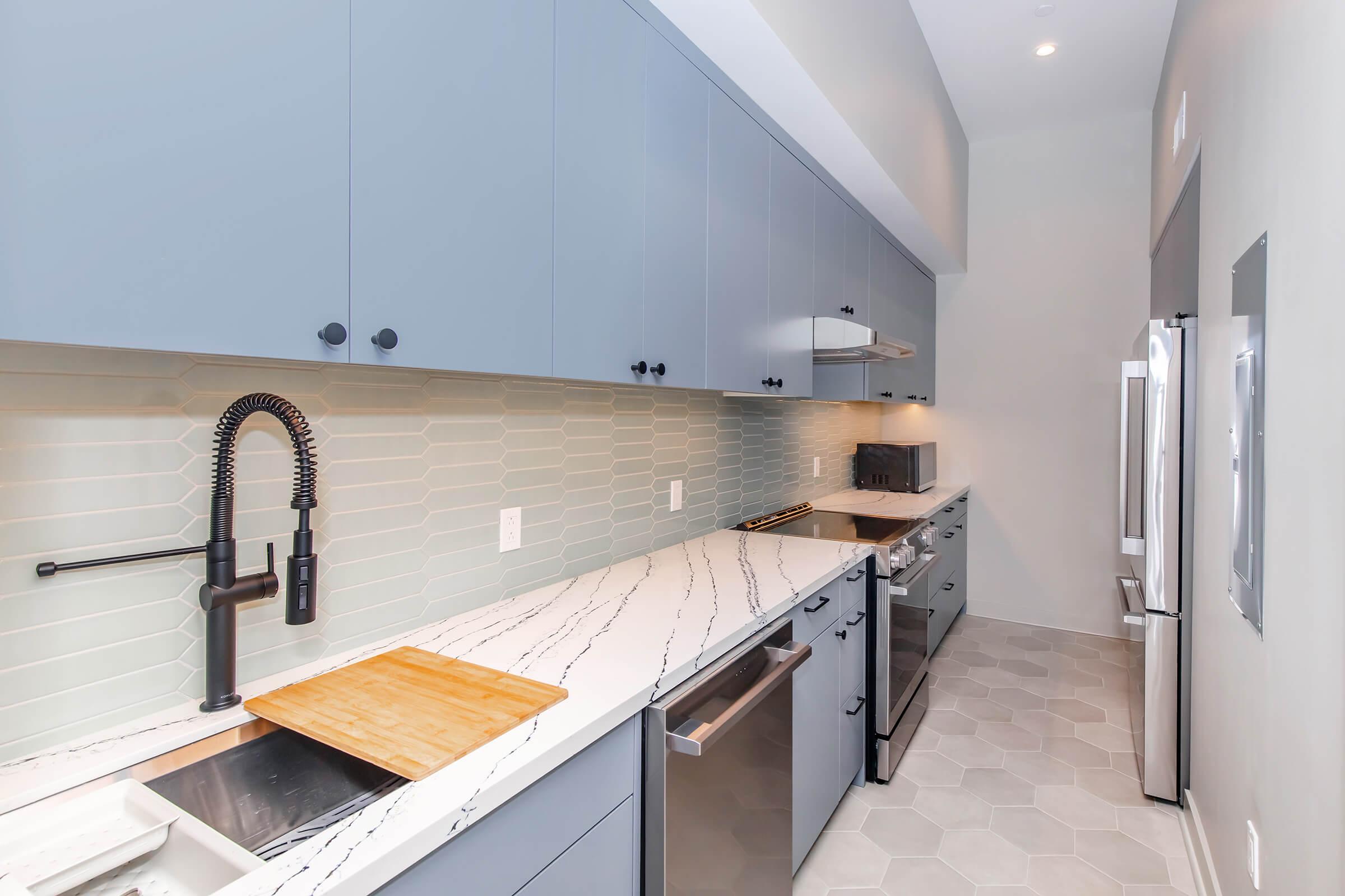 Modern kitchen featuring light gray cabinets, a white marble countertop with a wood cutting board, and a black faucet. The backsplash consists of light green hexagonal tiles, and there are stainless steel appliances, including an oven, microwave, and refrigerator, creating a sleek, contemporary aesthetic.