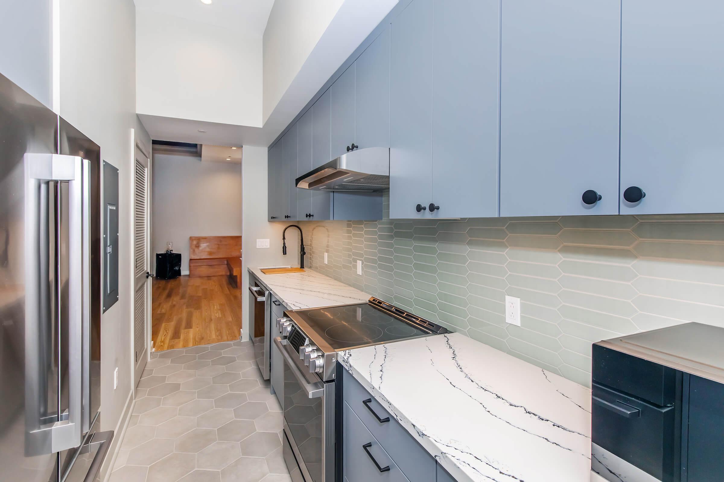 Modern kitchen featuring light blue cabinets, a stainless steel refrigerator, and a marble countertop. The backsplash is composed of hexagonal tiles, and there is a black faucet above the sink. The kitchen has wood flooring and a glimpse of an adjacent room in the background.