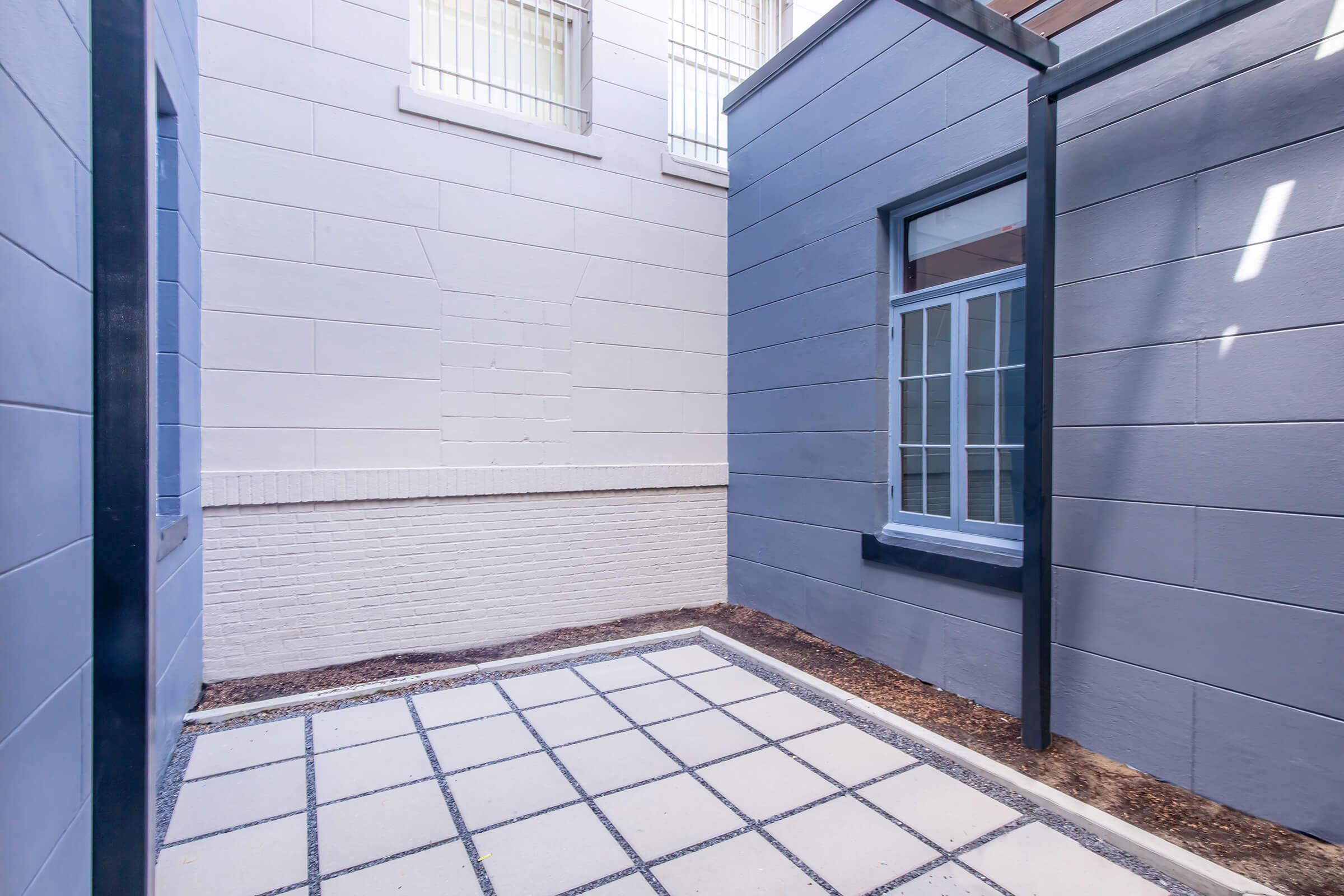 A view of a narrow courtyard featuring two grey walls, one flat and one with a window. The ground is paved with white stones arranged in a grid pattern. Light filters in from a window above, creating a bright and open atmosphere despite the enclosed space.