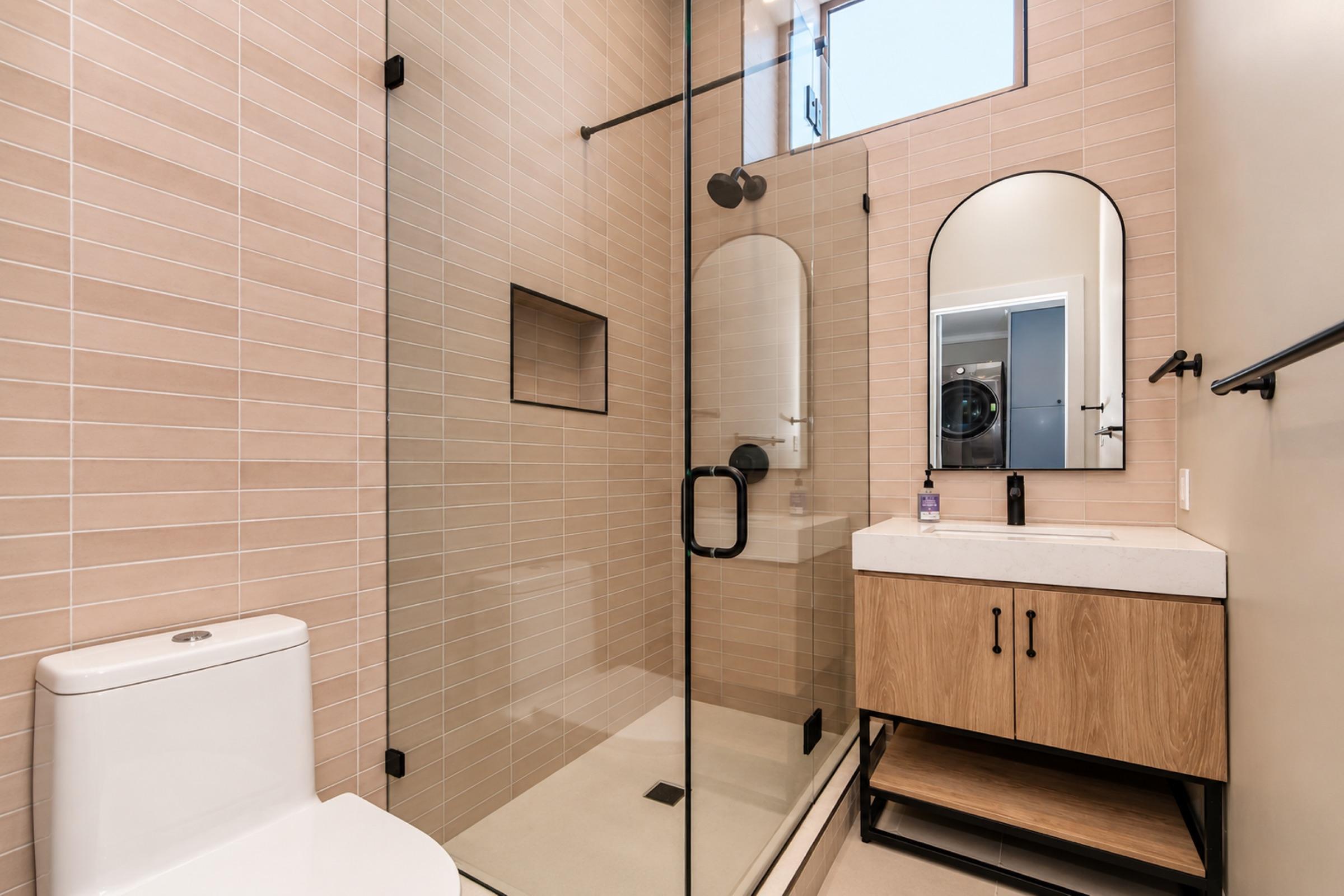 Modern bathroom featuring a glass-enclosed shower with beige tiled walls, a circular mirror above a wood vanity with a white countertop, and a sleek toilet. A window allows natural light, enhancing the clean and contemporary design.