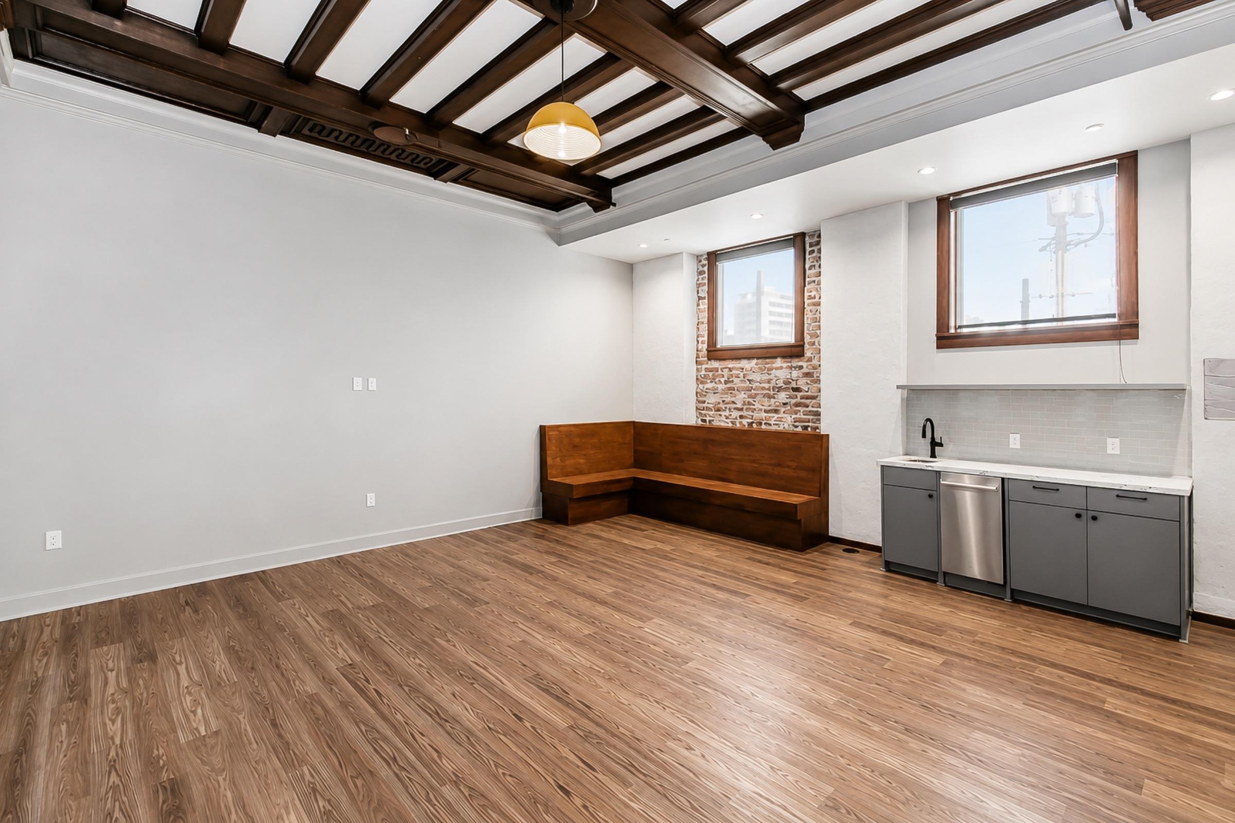 A spacious, modern room with wooden flooring, featuring exposed brick walls, a cozy built-in wooden bench, and a kitchen area with stainless steel appliances. Large windows allow natural light to fill the space, complemented by a stylish ceiling with wooden beams.