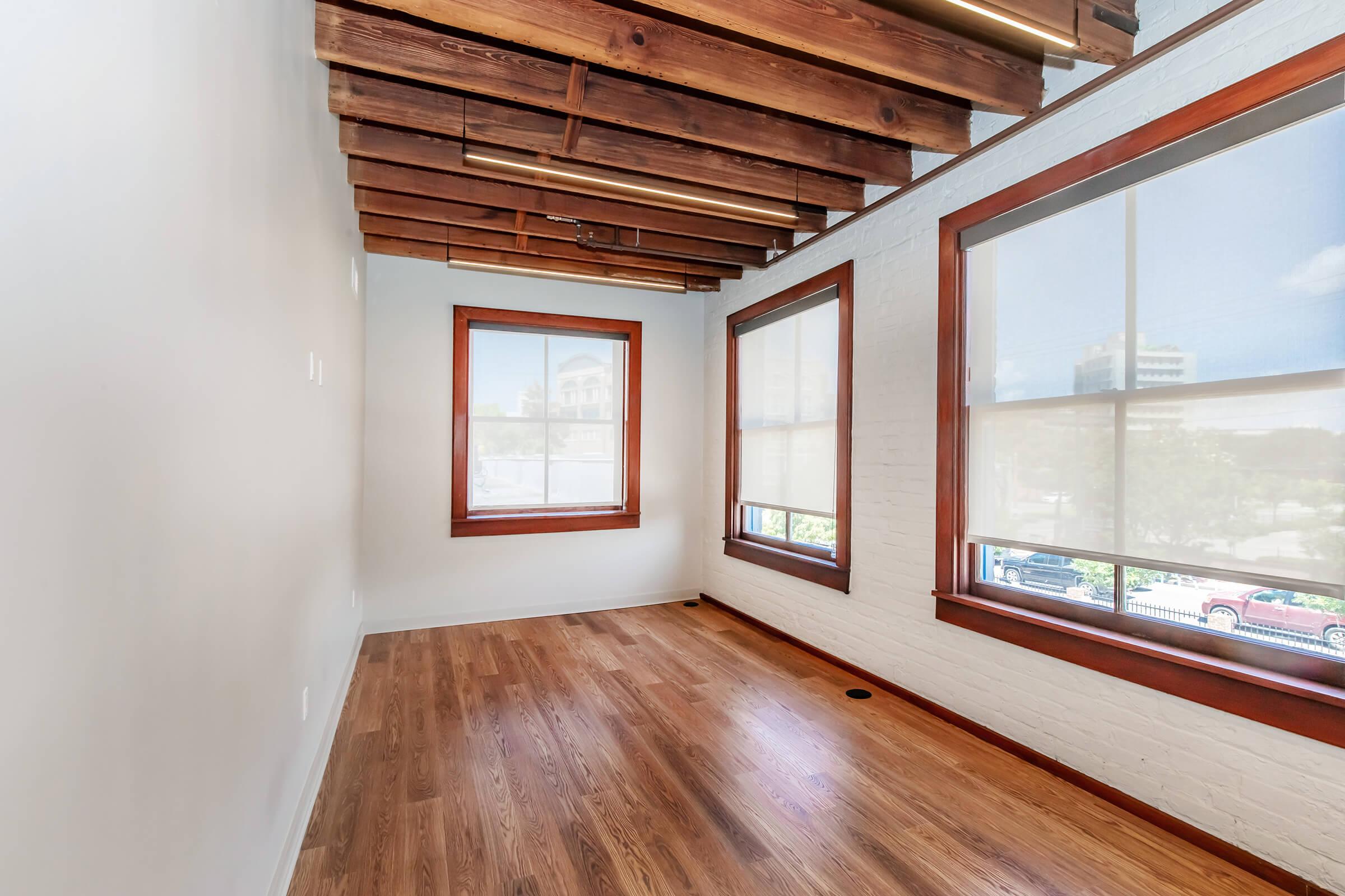 Empty room featuring wooden beams on the ceiling, large windows with blinds, and hardwood flooring. The white brick walls add a bright and modern feel. Natural light flows in through the windows, illuminating the space, which is suitable for various purposes such as an office or living area.