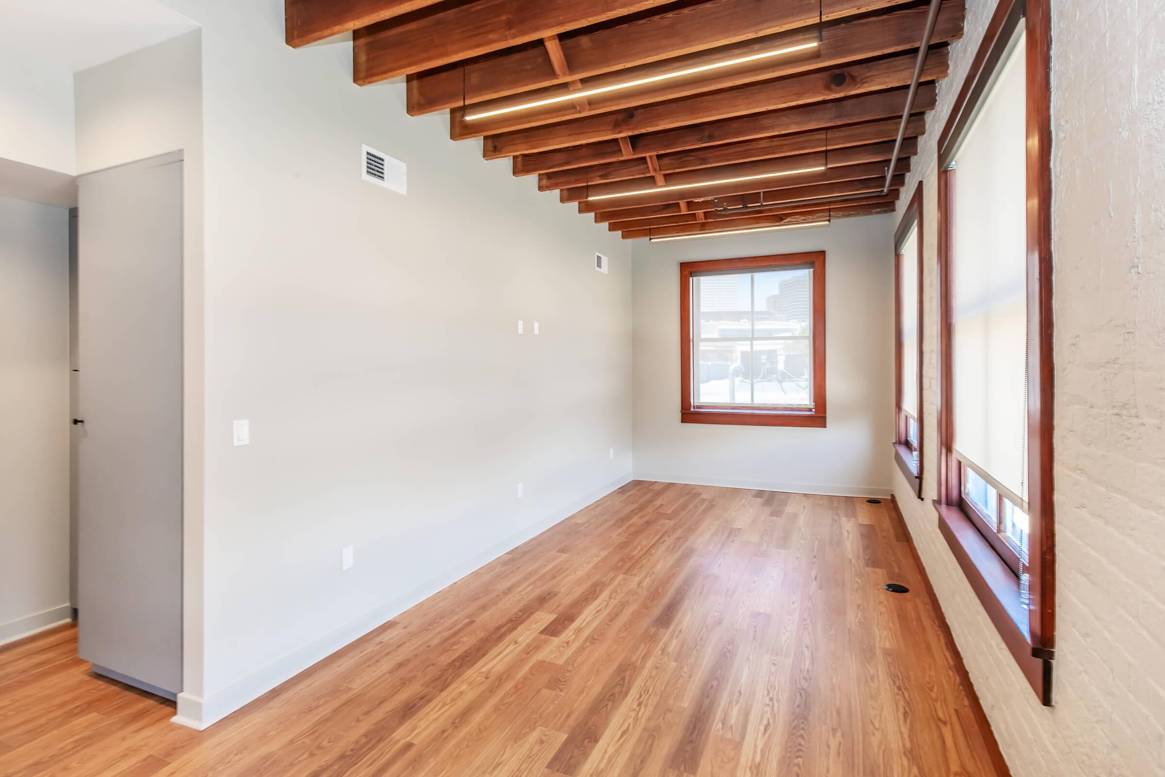 A well-lit room featuring wooden beams on the ceiling, hardwood flooring, and large windows with shades. The walls are painted light gray, creating a clean and modern atmosphere. There is a door on the left leading to another space, and the room is empty, suggesting it is ready for furnishing.