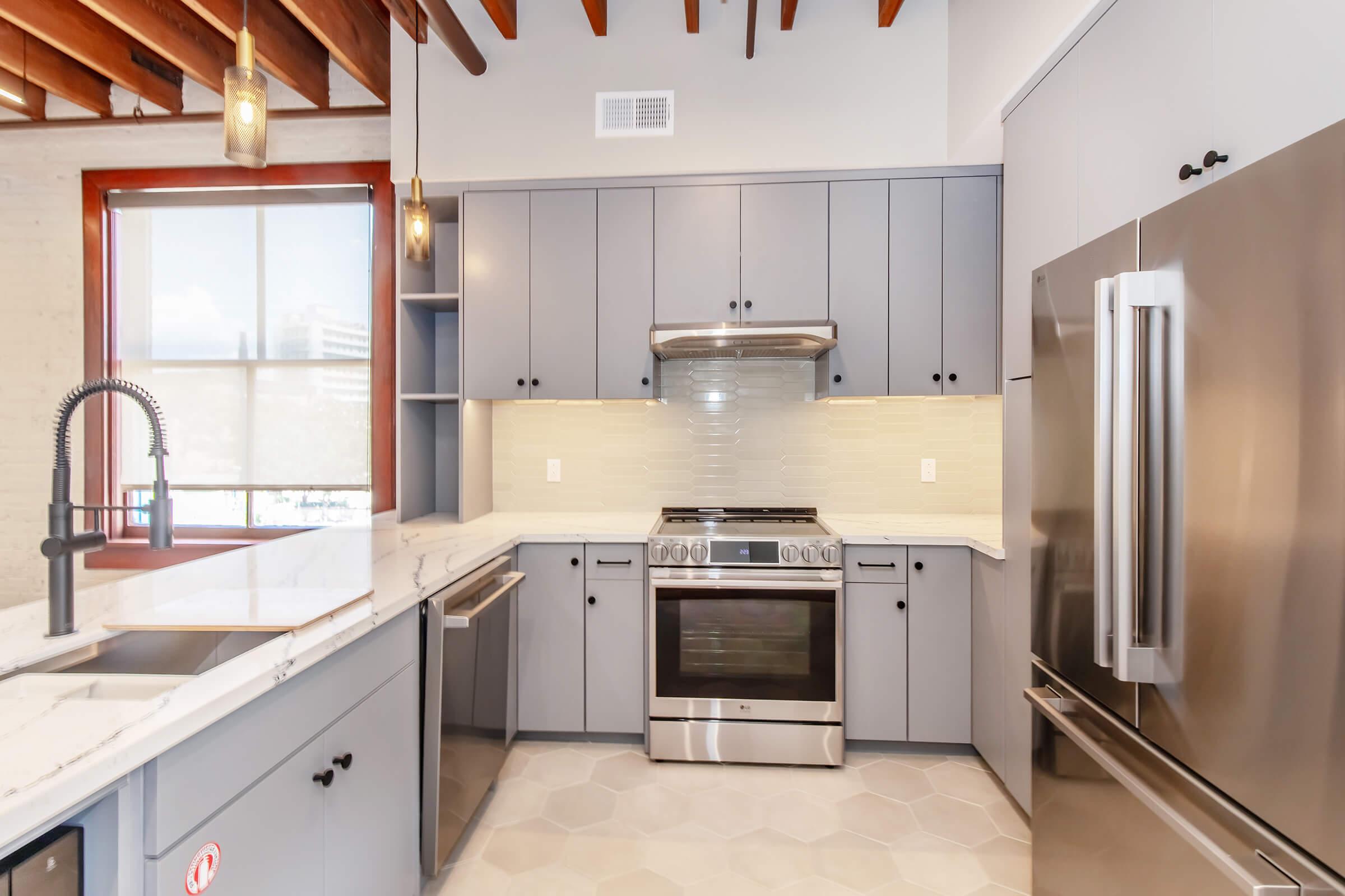 A modern kitchen with gray cabinetry, stainless steel appliances, and a white marble countertop. The kitchen features a gas stove, a double-door refrigerator, and a large sink with a sleek faucet. Natural light streams in through a large window, highlighting the exposed wooden ceiling beams.