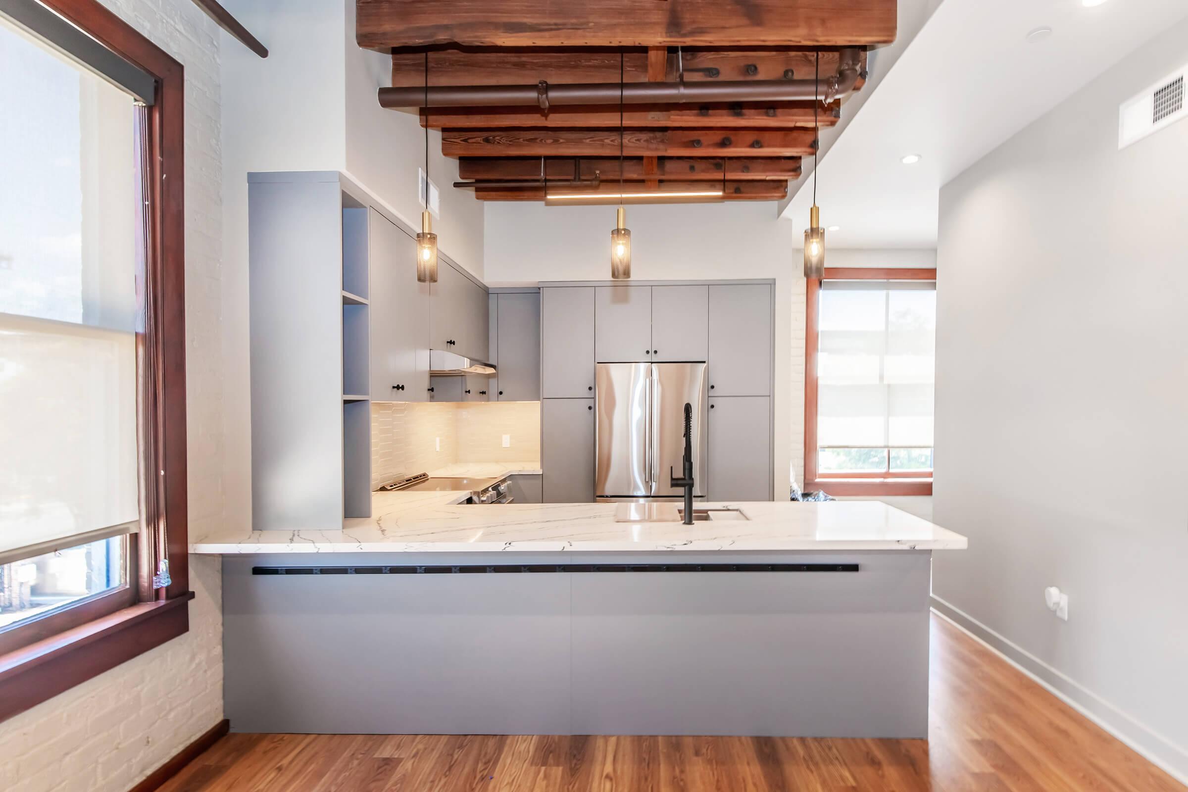 Modern kitchen featuring a central island with a white marble countertop, stainless steel appliances, gray cabinetry, and wooden beams on the ceiling. Large windows provide natural light, highlighting the warm wooden flooring and contemporary design elements.