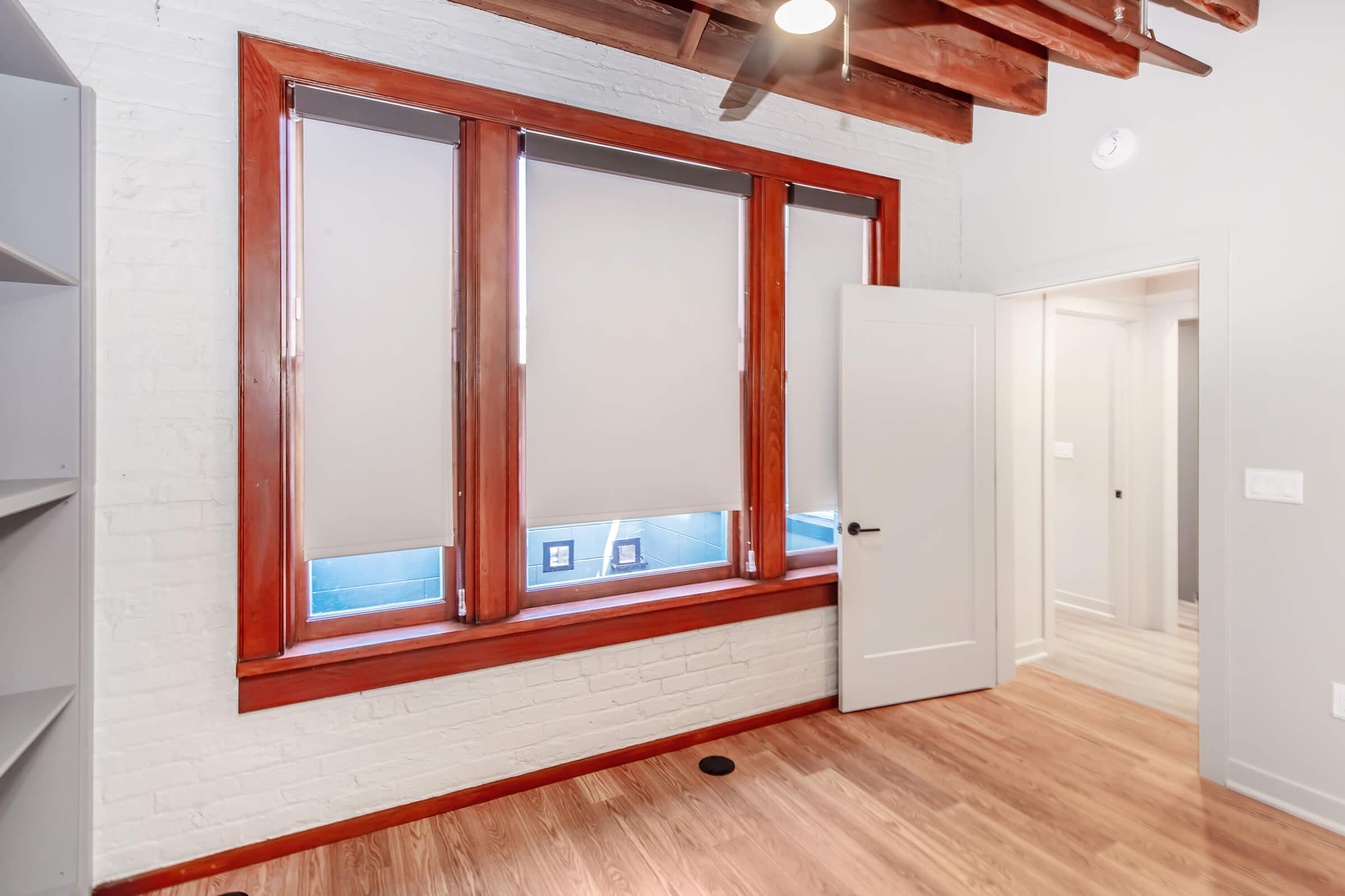 Interior view of a room featuring exposed wooden beams, large windows with blinds, and a door leading to another part of the space. The walls are painted white with a light wooden floor. A bookshelf is visible on the left side, enhancing the modern, airy feel of the area.