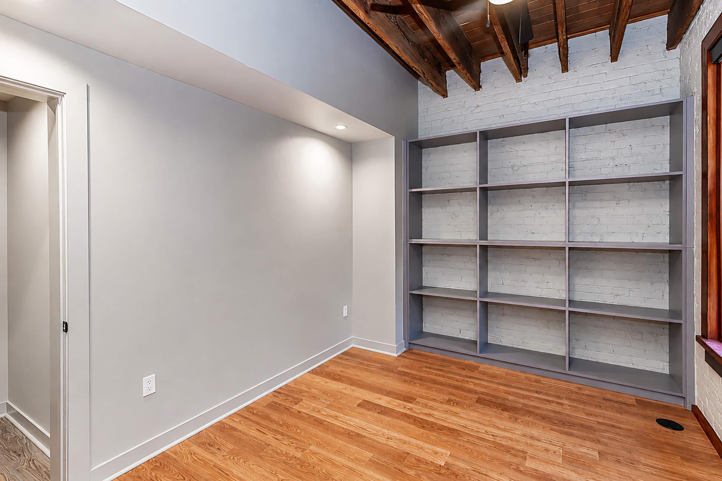 Interior of a modern room featuring gray walls, a wooden ceiling with exposed beams, and a built-in bookshelf against a white brick wall. The floor is made of light hardwood, and there is a doorway visible on one side. Natural light comes from a window, enhancing the spacious feel of the room.
