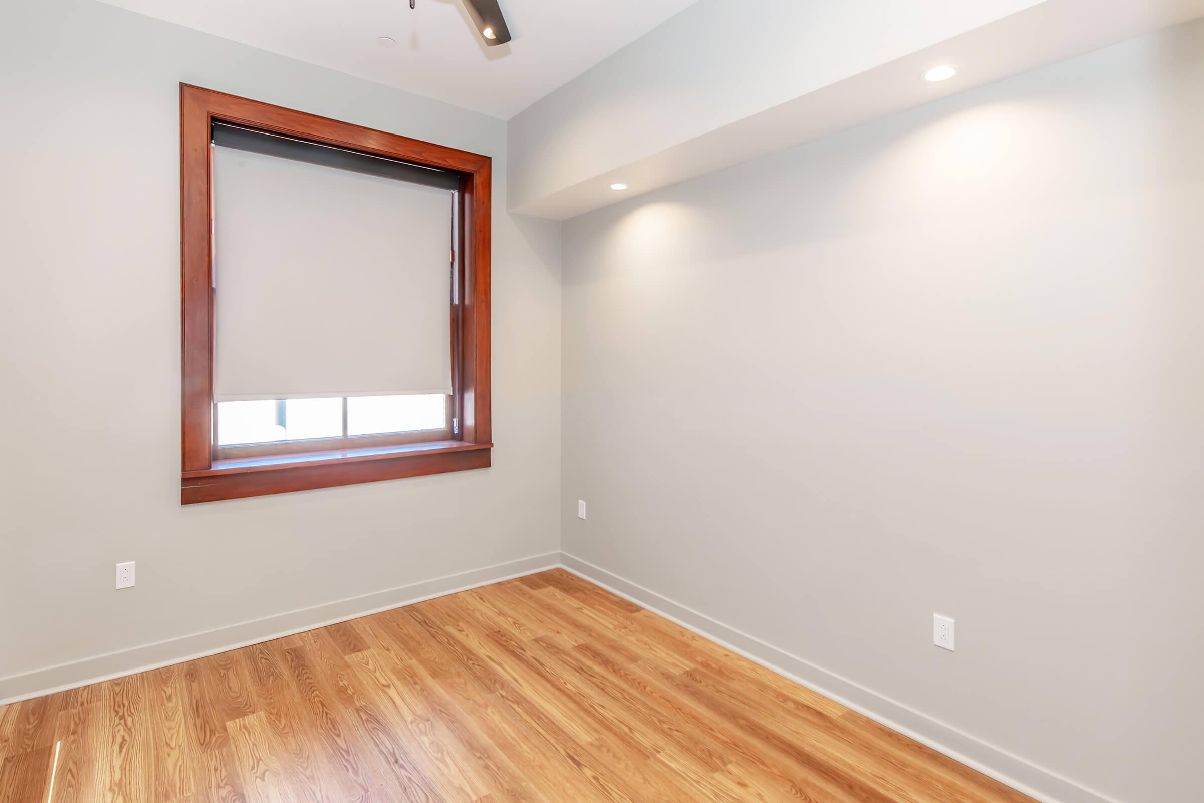 A minimalistic empty room featuring a large window with a rolled-up blind, wooden trim, and hardwood flooring. The walls are painted in a light gray color, with soft lighting from recessed fixtures illuminating the space.