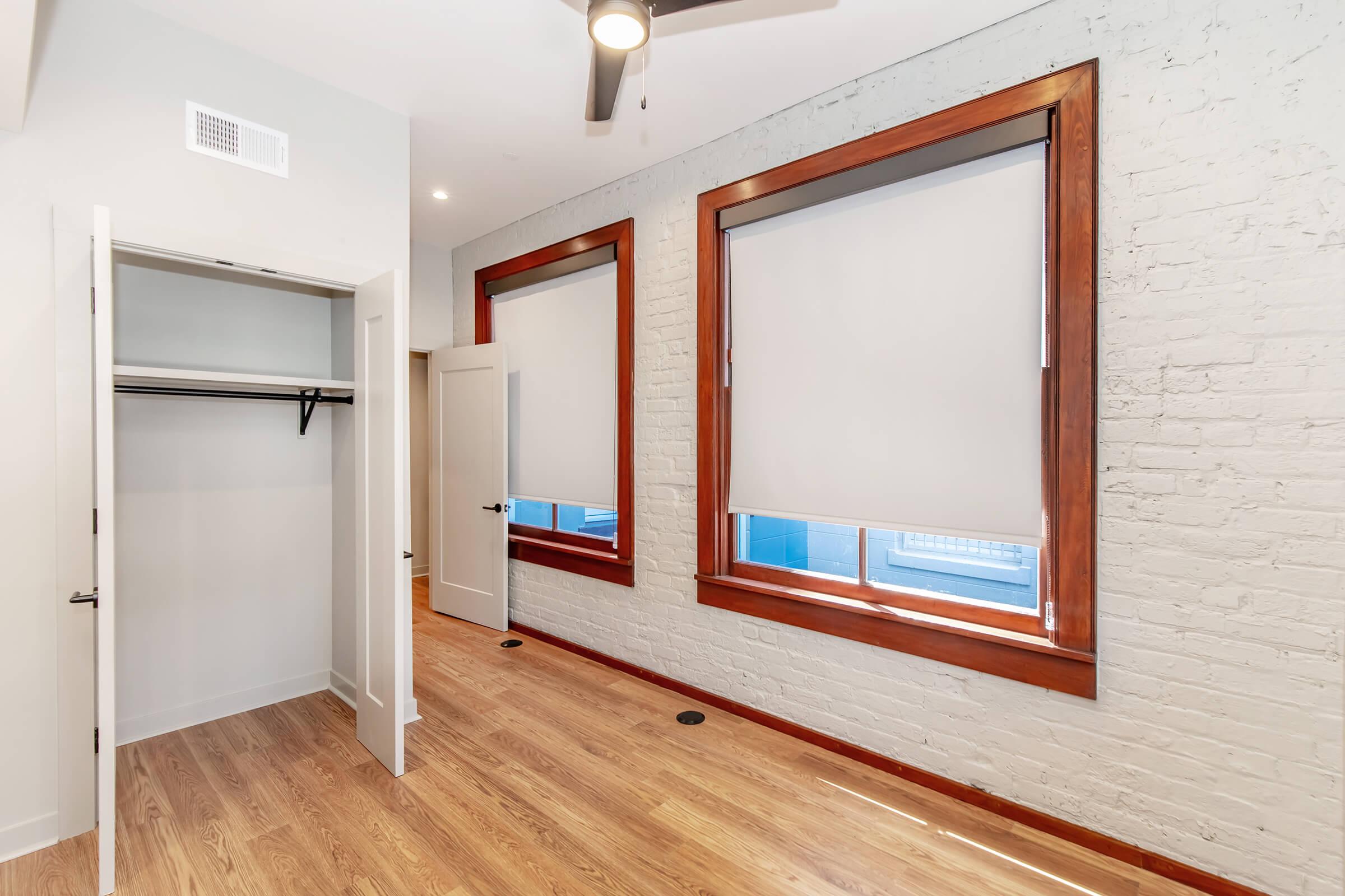 A modern room featuring light wood flooring, exposed brick walls, and two large windows with roller shades. A closet with a hanging rod is visible to the left, and the room is illuminated with natural light. The overall design has a minimalist and contemporary feel.