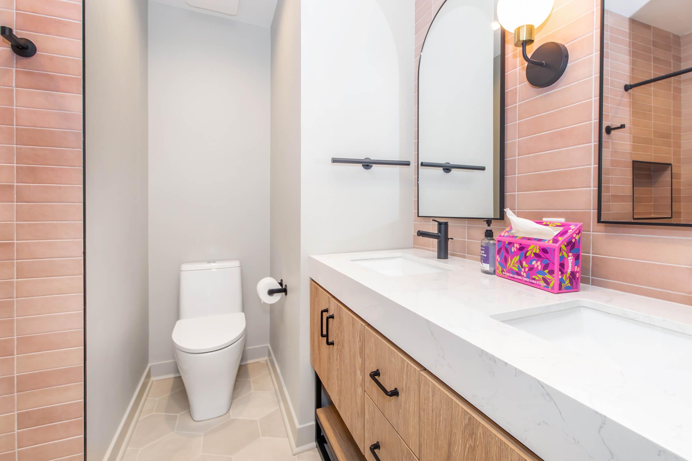 A modern bathroom featuring a clean design with light pink tiled walls, a white toilet, and a double vanity with a marble countertop. Two round mirrors are mounted above the sinks, and a colorful tissue box adds a pop of color. The floor is made of light-colored hexagonal tiles, creating a fresh and contemporary look.