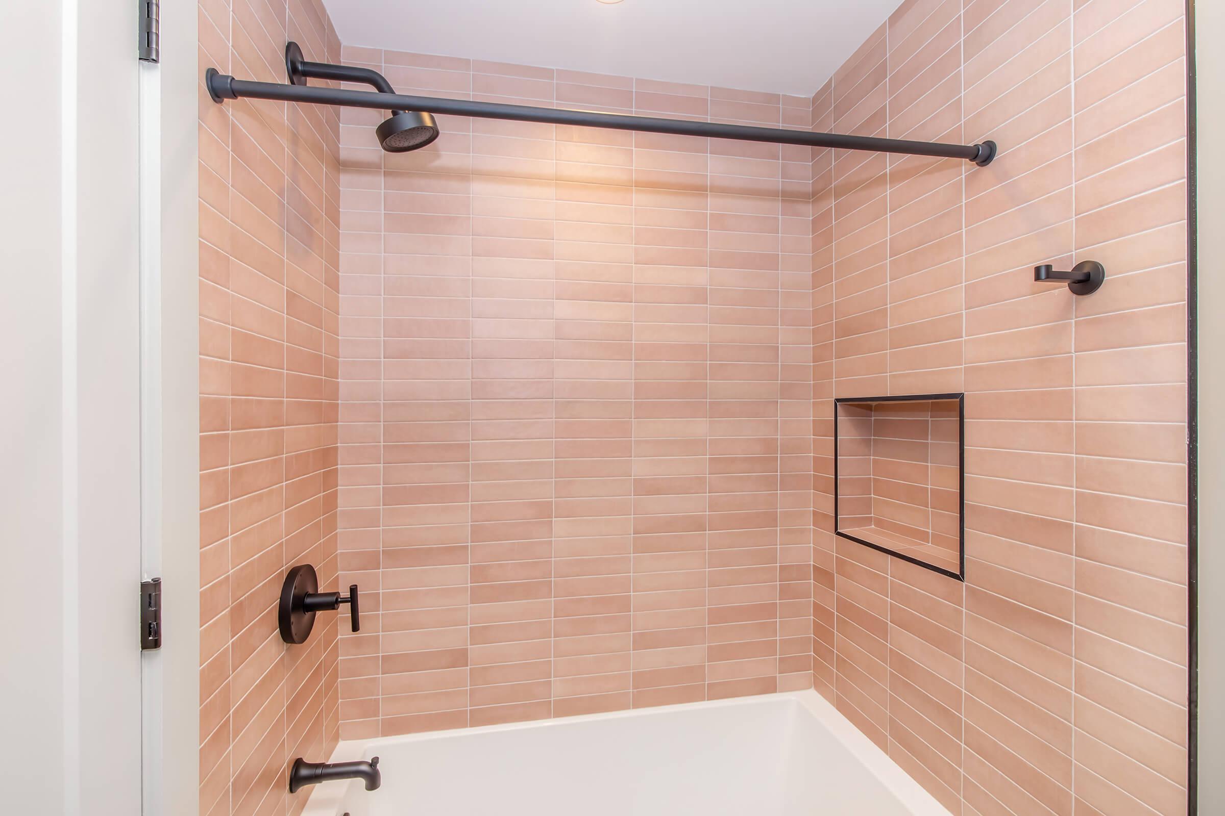 A modern bathroom shower featuring a warm-toned tiled wall with a sleek, black showerhead and pipe. There's a built-in niche for toiletries and a minimalist shower control handle. The overall design is clean and contemporary, emphasizing neutral colors and smooth lines.