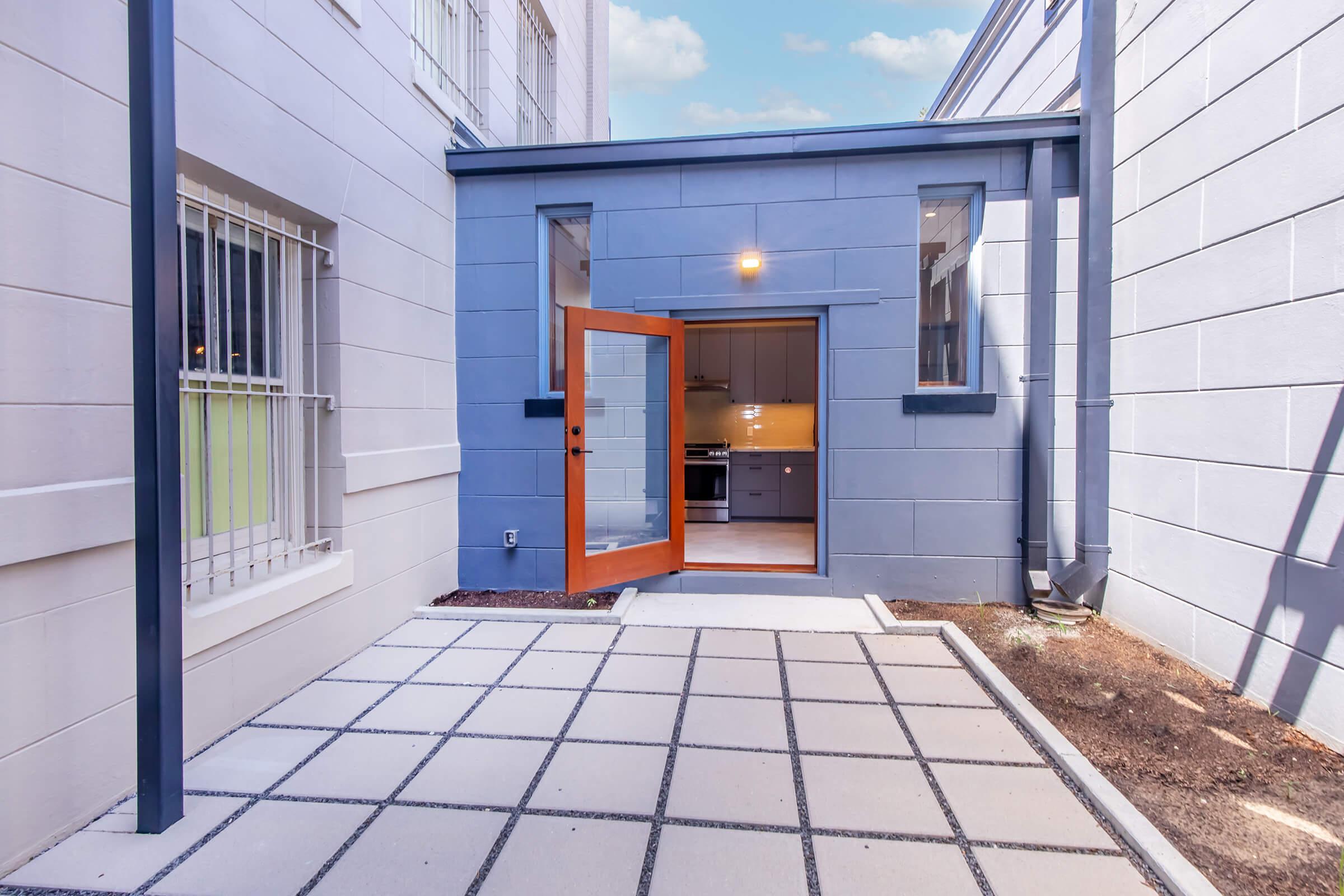 A view of a small outdoor patio area with a modern design. The space features a grey wall with large windows and an open door leading to a kitchen. Concrete tiles cover the ground, and there are minimal plants along the edges, creating a clean and inviting atmosphere.