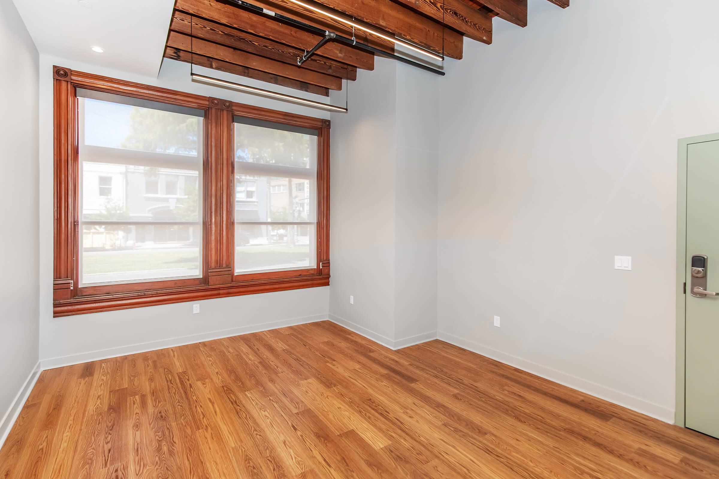 Bright, empty interior room with large windows showcasing wooden frames, high ceilings with exposed beams, and hardwood flooring. The walls are painted in a light color, and there is a green door on the right side. Natural light fills the space, creating an inviting atmosphere.