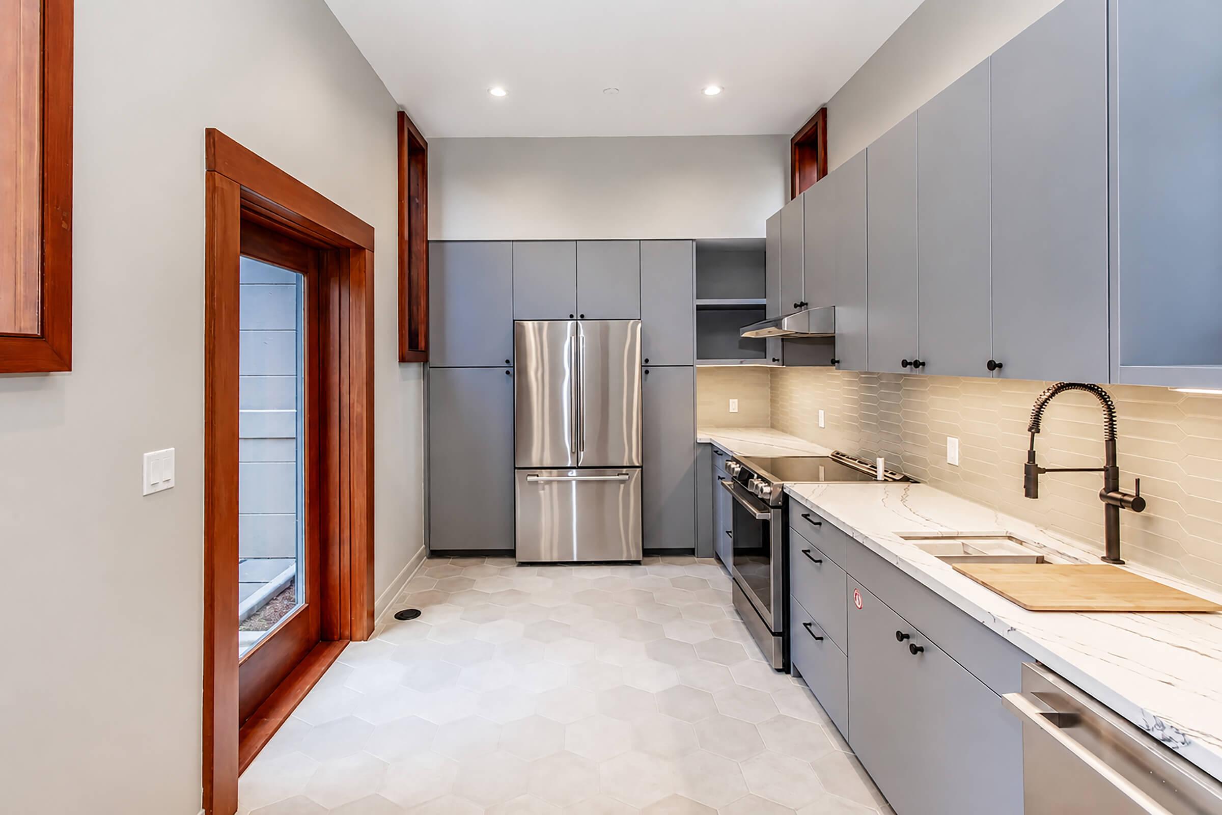 Modern kitchen featuring gray cabinetry, stainless steel appliances including a refrigerator and stove, and a light-colored hexagonal tile floor. A large window is present, providing natural light, and there are wooden accents around the doorway. The space is well-organized and stylishly designed.