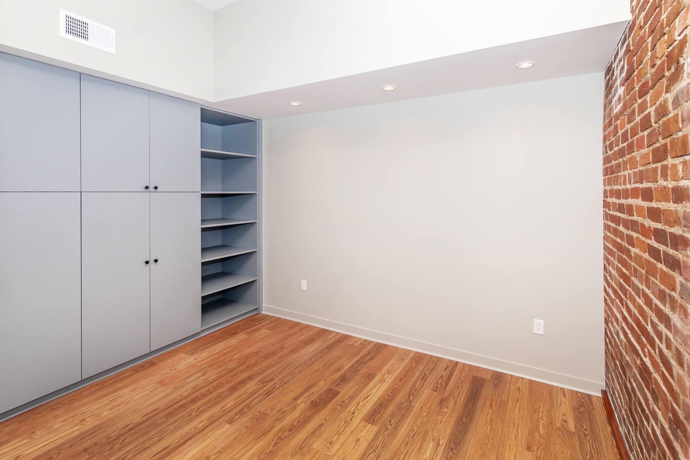 A minimalist room featuring a brick accent wall, a light-colored wall, and built-in cabinetry in gray. The hardwood floor adds warmth to the space. There are recessed lights on the ceiling, and one corner has open shelving. The overall design is modern and uncluttered, suitable for various uses.