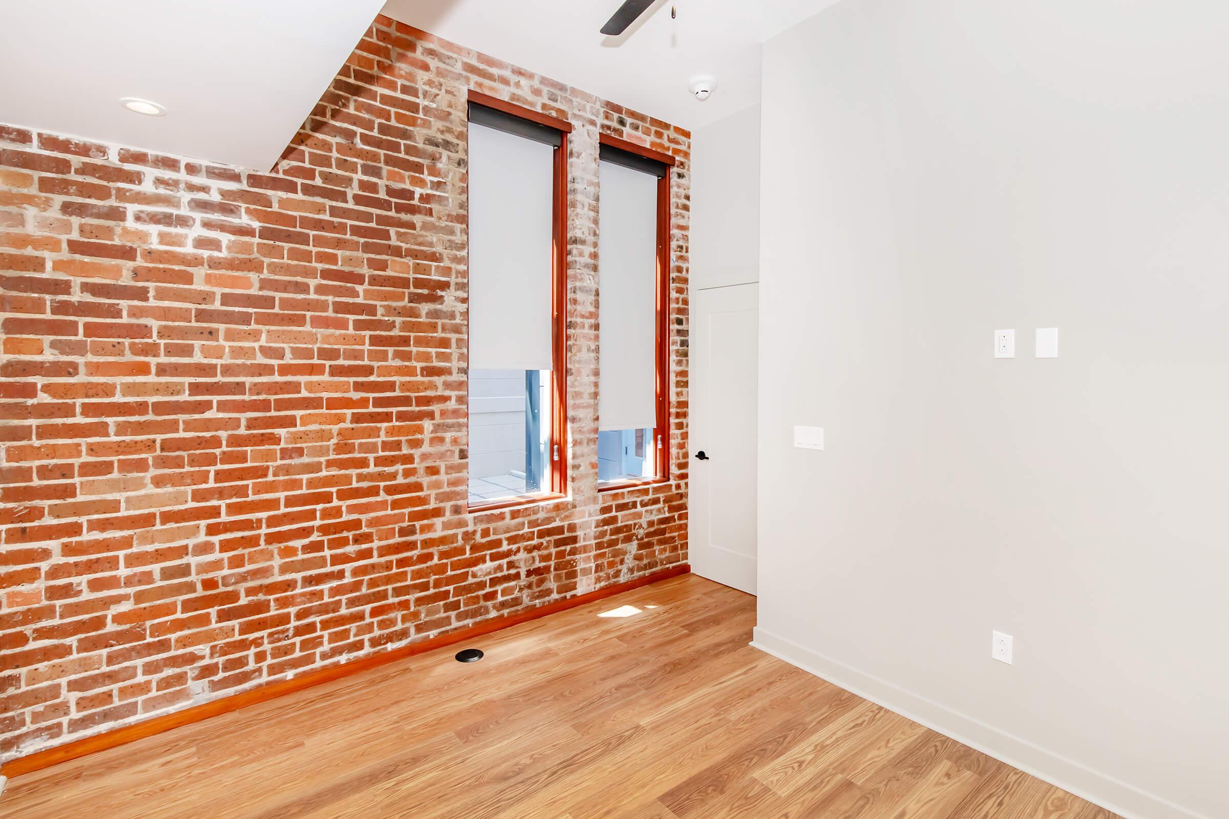 A modern interior featuring a textured brick wall, large windows with roller shades, and a light-colored room with wooden flooring. The space is well-lit with natural light, creating a cozy atmosphere.