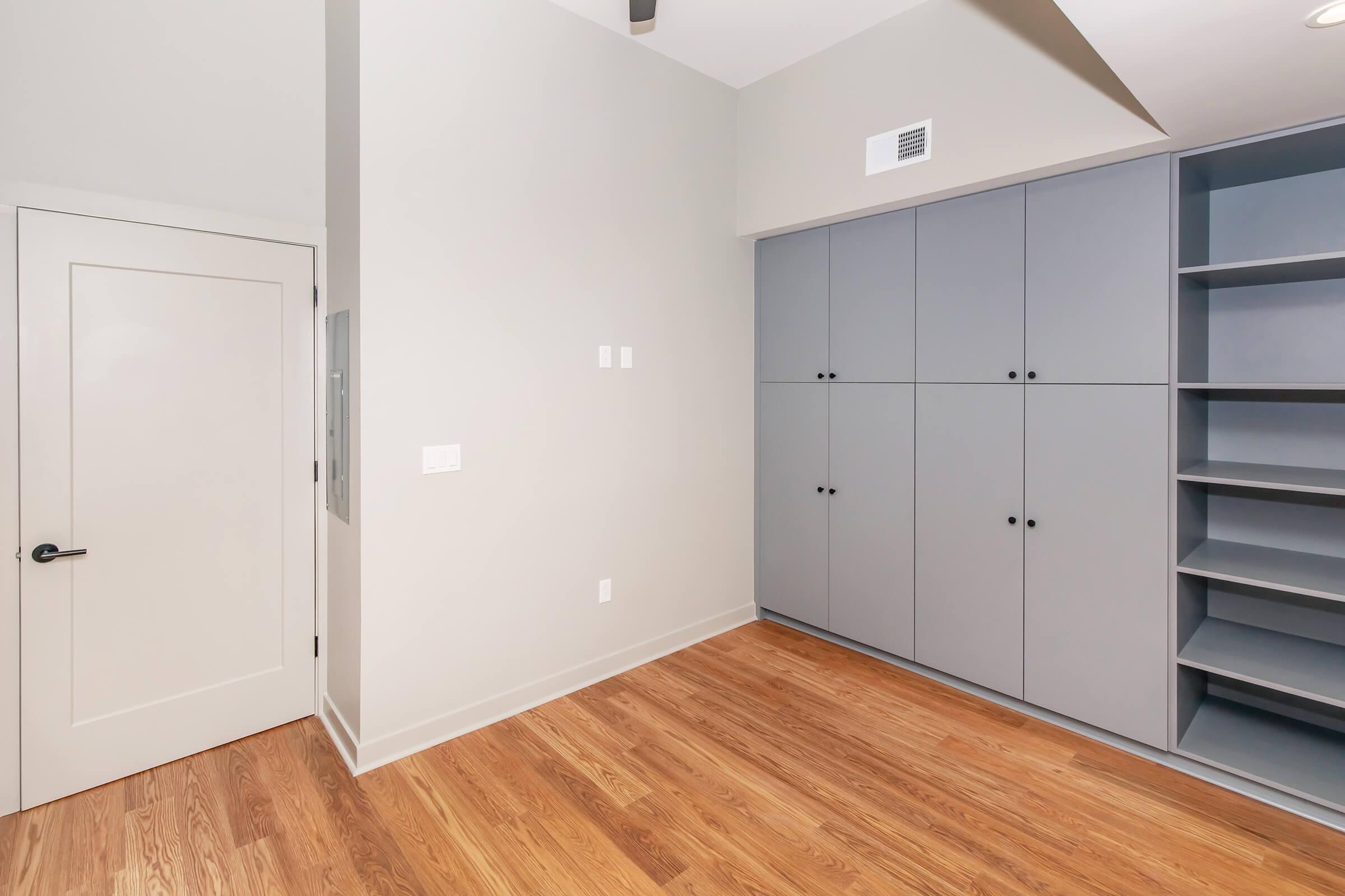 A spacious interior room featuring light gray walls and a wooden floor. On one side, there is a closed white door, and along another wall, built-in gray cabinets with shelves. The room has a modern and minimalistic design, with a ceiling fan visible.