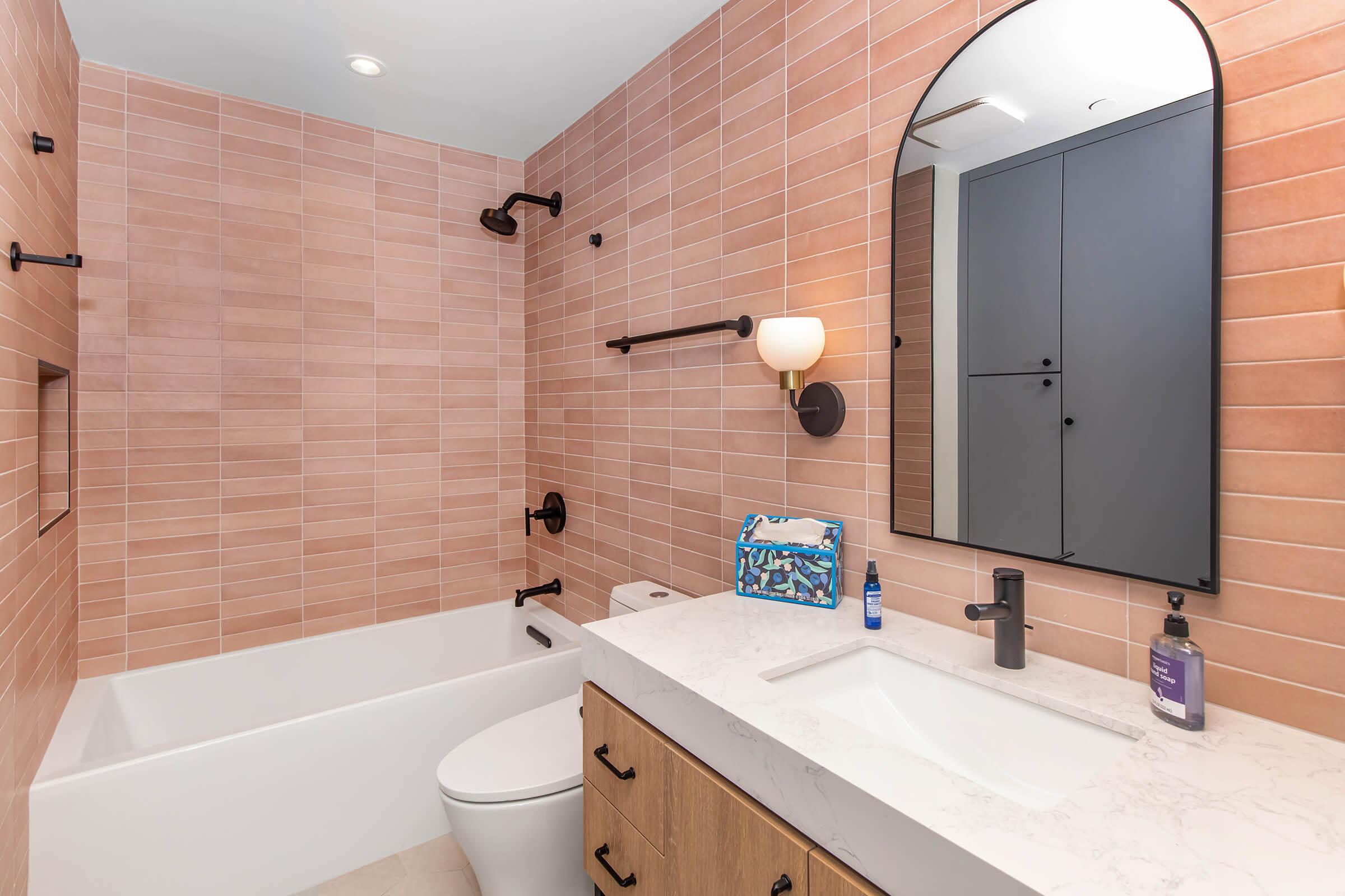 A modern bathroom featuring a bathtub with a shower, pink tiled walls, a marble countertop with a sink, a wall-mounted mirror with an arched top, and stylish fixtures. The space includes a light fixture, black towel bars, and organized toiletries on the countertop.
