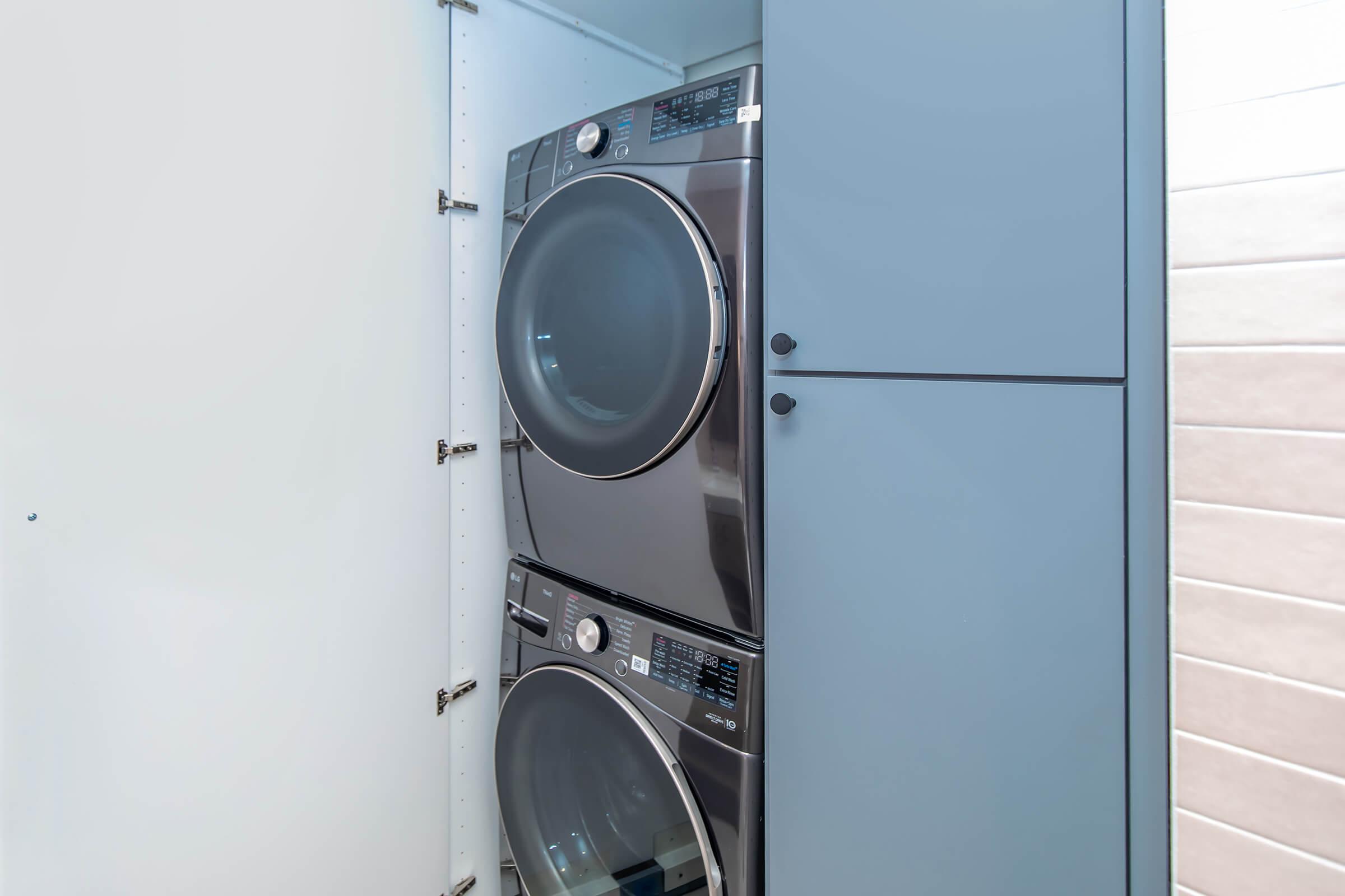 A modern laundry setup featuring a stacked washer and dryer in a stylish, light-colored laundry room. The appliances are sleek and front-loading, with a nearby cabinet offering additional storage space. The walls are neutral, contributing to a clean and organized appearance.