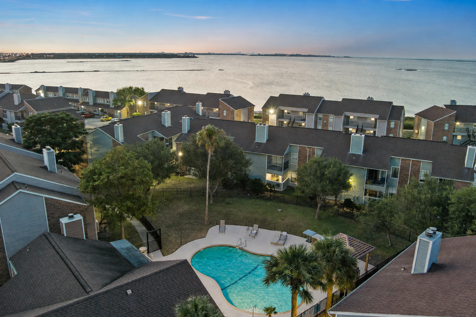 Aerial view of an apartment complex near a body of water at dusk. The foreground features a swimming pool surrounded by palm trees, with well-maintained lawns. Multiple residential buildings line the background, leading toward the water's edge, reflecting the evening sky's soft colors.