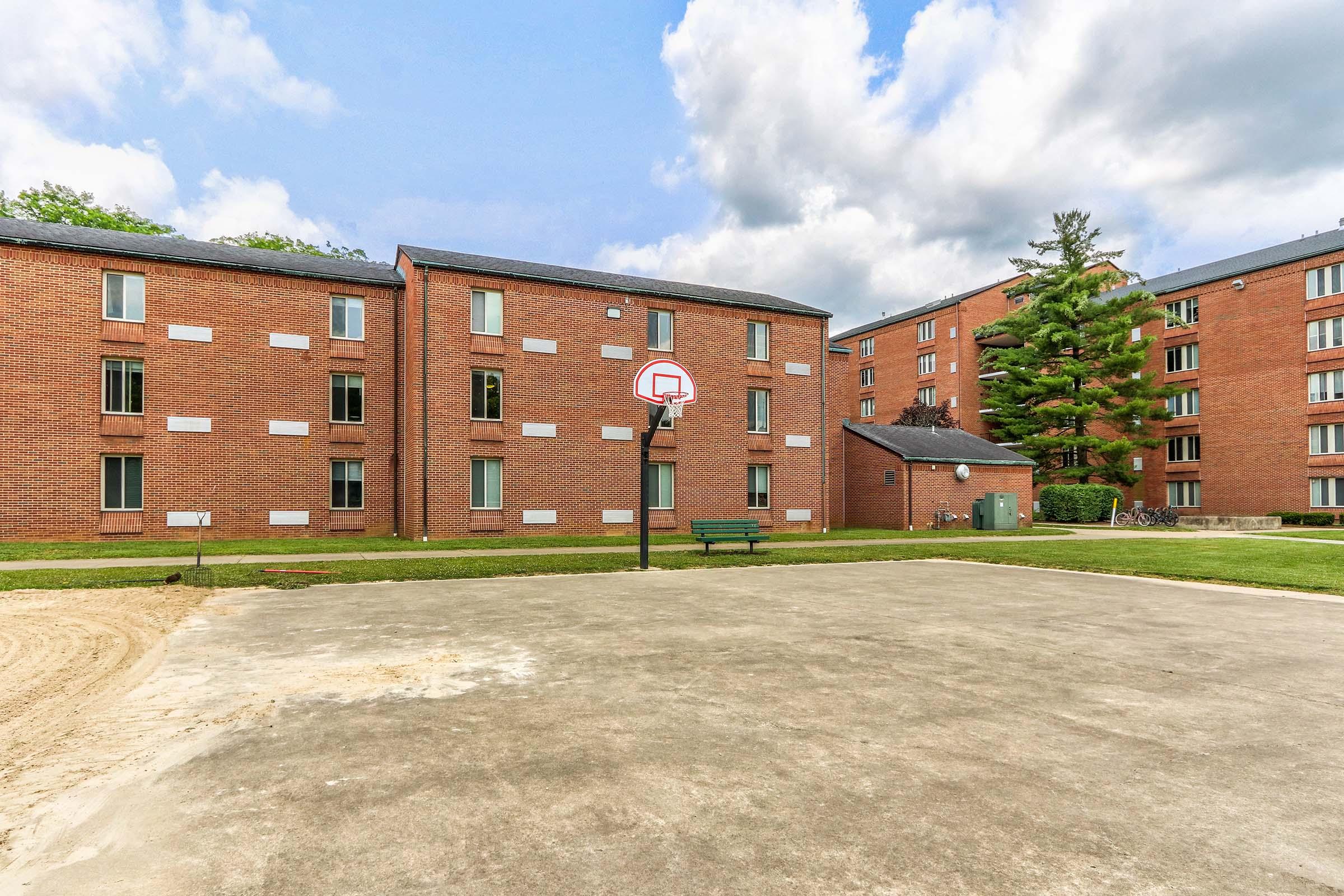 A basketball court in front of a brick building complex. The court features a basketball hoop and a bench nearby, with a sandy area around the court. There are trees in the background and a cloudy sky overhead, creating a calm outdoor environment.