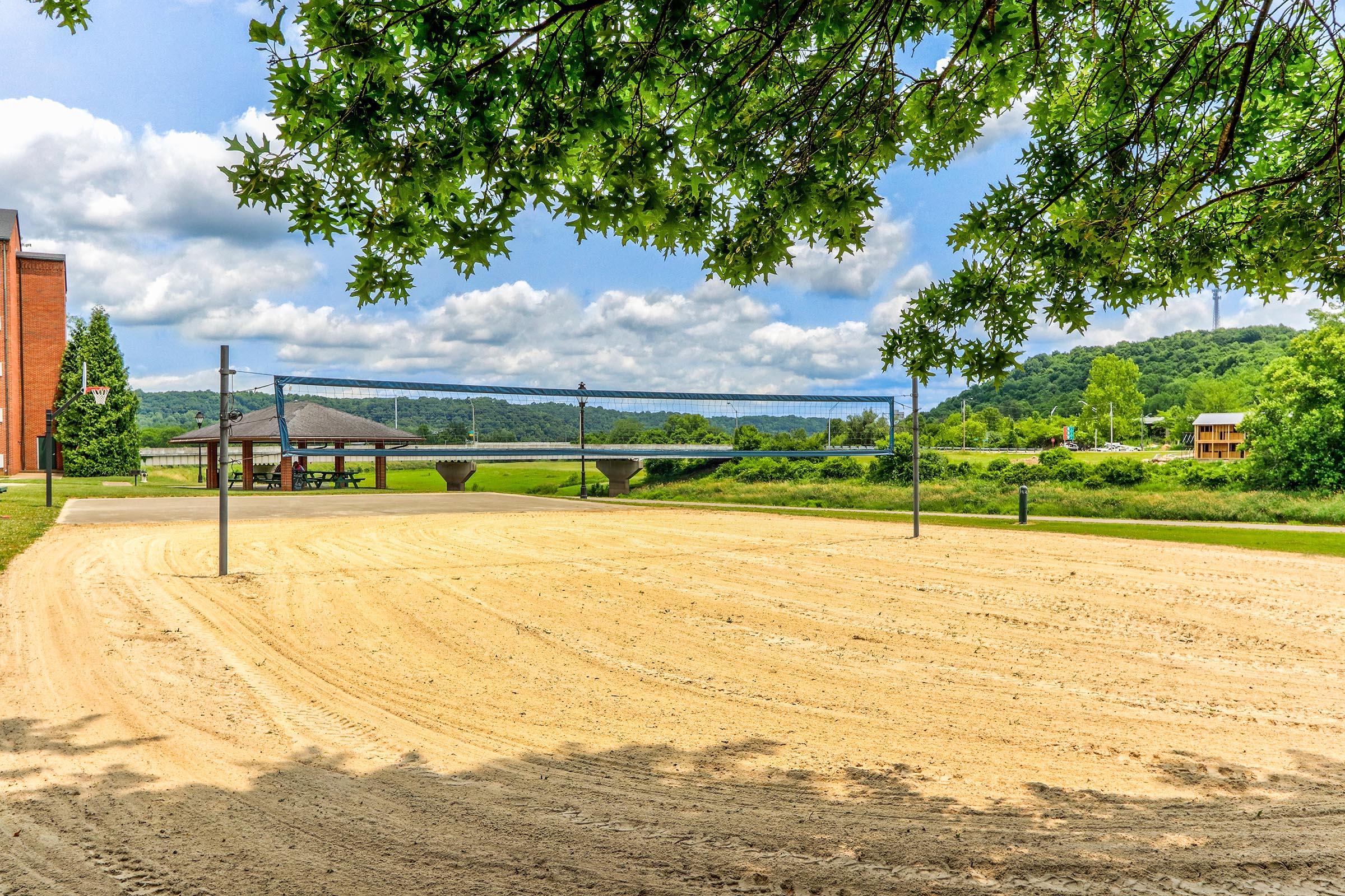 A sandy volleyball court set against a backdrop of green hills and a blue sky with fluffy clouds. Trees frame the scene, adding a natural touch. Nearby, a gazebo is visible, along with a glimpse of a building and some distant structures. The area looks peaceful and inviting, perfect for outdoor activities.