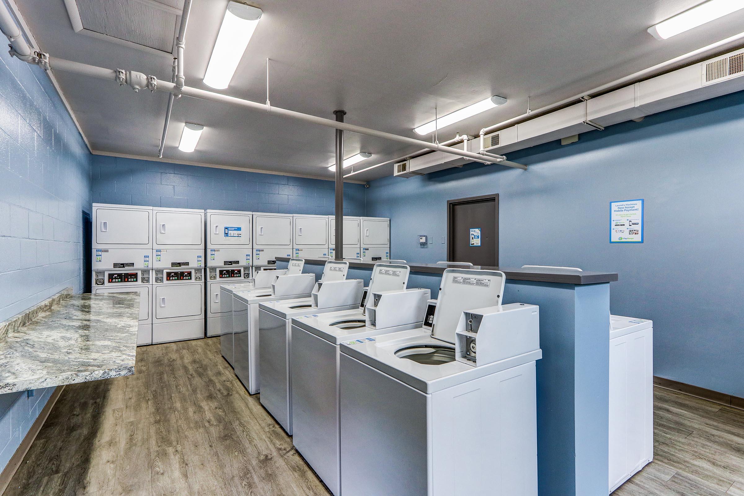 A clean and modern laundry room featuring several rows of washing machines and dryers. There is a countertop area for folding clothes, and the walls are painted in a light blue color. The space is well-lit with fluorescent lights, creating a bright and inviting atmosphere.