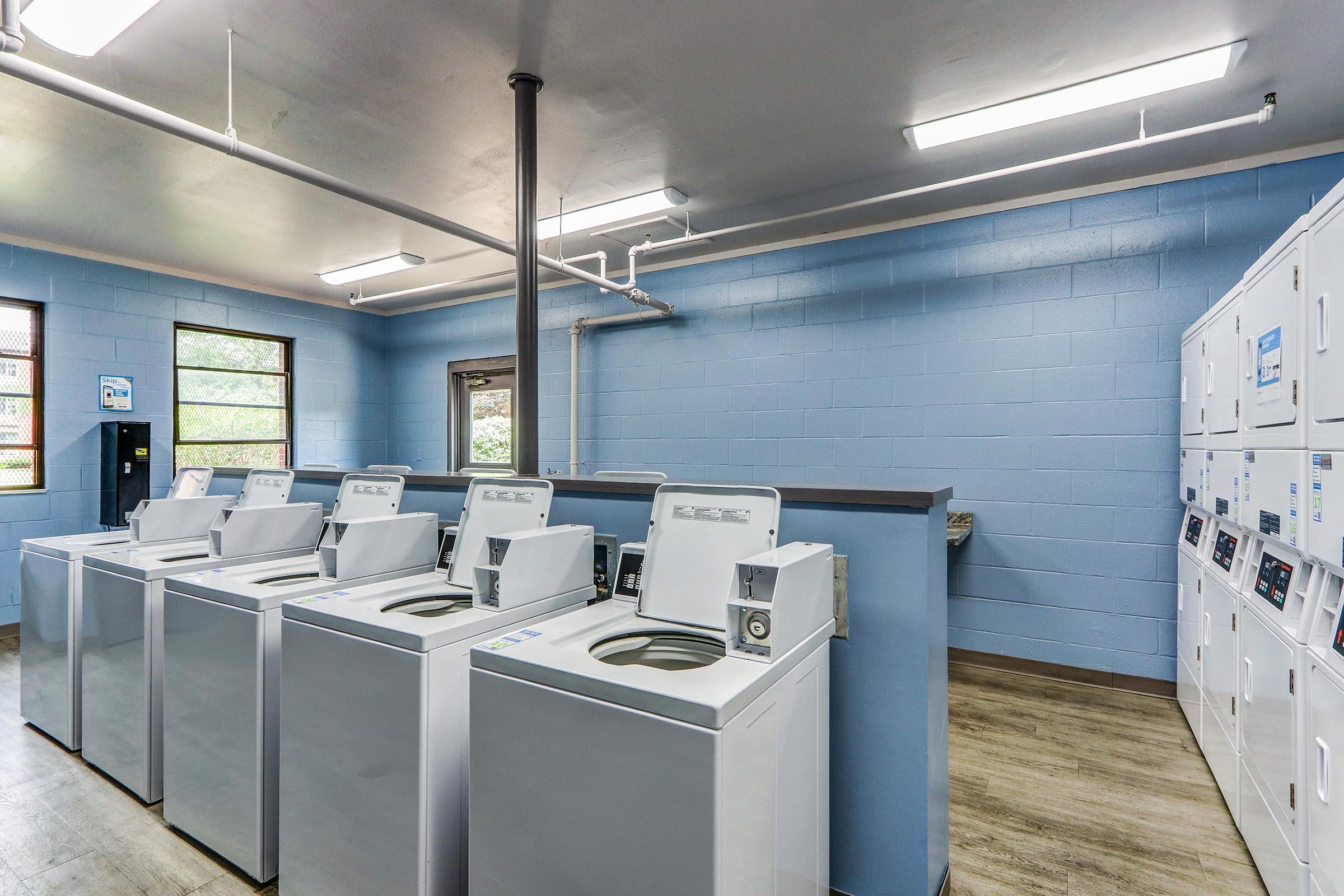 A clean, bright laundry room with several white washing machines lined up against a blue wall. There's a row of white dryers and a payment machine nearby. The space has windows allowing natural light, and the flooring is a light wooden finish. The overall setting appears organized and well-maintained.